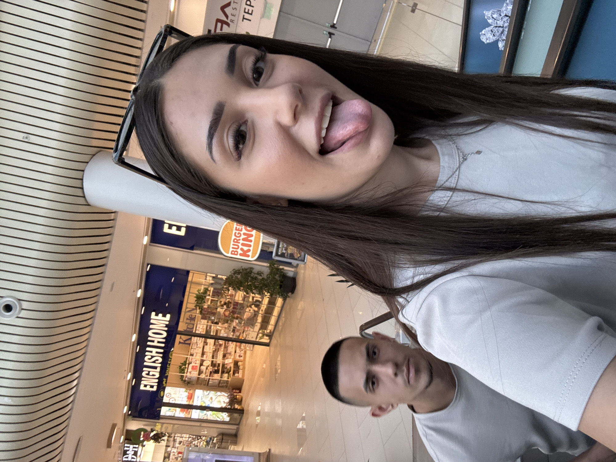 The image captures an indoor scene within a shopping mall, located in Plovdiv, Bulgaria. The perspective suggests a selfie taken by the female in the foreground.
The foreground is dominated by a young adult female, positioned slightly to the right. She has long, straight, dark brown hair with a central parting. Black-framed sunglasses rest on top of her head. Her mouth is open, and her tongue is extended, directed towards the camera, which she gazes into directly. She wears a light grey, short-sleeved, crew-neck t-shirt and a thin silver-colored chain necklace with a small pendant.
In the mid-ground, to the left of the female, a young adult male is partially visible from the chest up. He has short, dark hair, specifically a buzz cut, and faint stubble on his chin. He also wears a light grey, short-sleeved, crew-neck t-shirt and looks directly into the camera with a neutral expression. An escalator handrail is visible between the two individuals.
The background consists of a multi-level shopping mall environment. A prominent blue sign for "ENGLISH HOME" is visible above a retail store, which displays various home decor items. To the right, a circular orange sign for "BURGER KING" is partially visible. Further to the right, parts of a white sign bearing the Cyrillic letters "ГА" and "ТЕРА" are discernible, indicating a larger mall name such as "ГАЛЕРИЯ" (Galleria). The mall features a distinctive ceiling with numerous parallel dark lines on a light surface, large white support pillars, and light-colored tiled flooring. The scene is well-lit by internal artificial lighting.