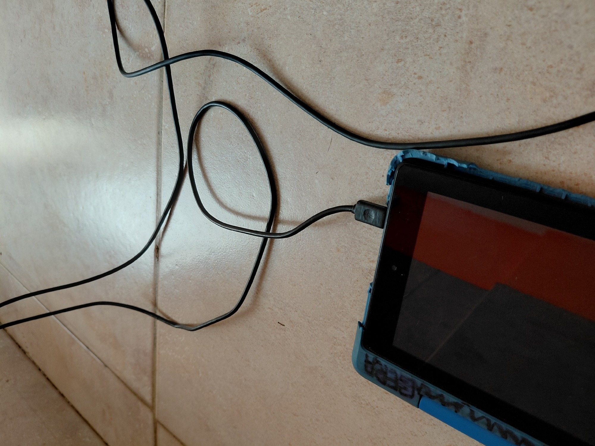 An overhead shot depicts a black tablet, fitted with a textured blue protective case, resting on a light-colored, tiled floor. A black charging cable is plugged into the tablet's left side, forming a large loop and extending upwards and out of the frame. The tablet's screen is dark, showing faint reflections, possibly of red and black tones. On the bottom edge of the blue protective case, partially obscured, is some black writing that appears to spell "GEBRA" or "CEBRA". The floor consists of large, rectangular tiles with visible grout lines and a slightly mottled, beige or off-white coloration, indicative of an indoor setting. The scene is brightly and evenly lit, without strong shadows to suggest a specific time of day or weather conditions. No activity beyond the tablet charging is discernible, and no people are present.
