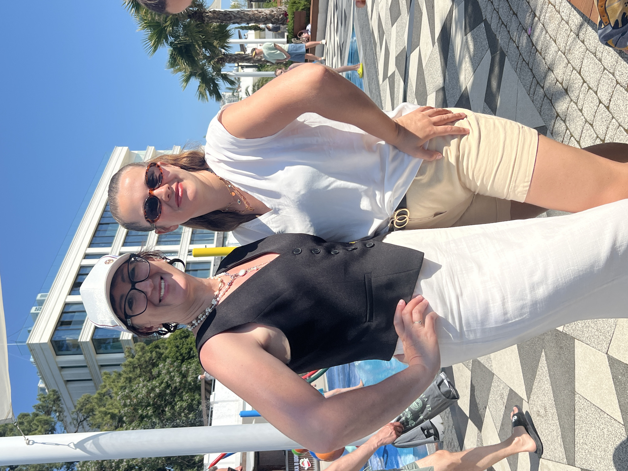 Two women stand side-by-side outdoors on a sunny day, posing for a photo. The woman on the left, wearing a white baseball cap, glasses, a black vest over a pearl necklace and a long white skirt, smiles at the camera. The woman on the right, with her hair pulled back, wears sunglasses, a white V-neck shirt, and light beige shorts. They are standing on a patterned paved area, with a swimming pool or water feature to their left and palm trees and a modern building in the background, suggesting a resort or hotel environment. The presence of palm trees and the specific architectural style of the building point to the city of Sochi, Russia, known for its subtropical climate and resort destinations.