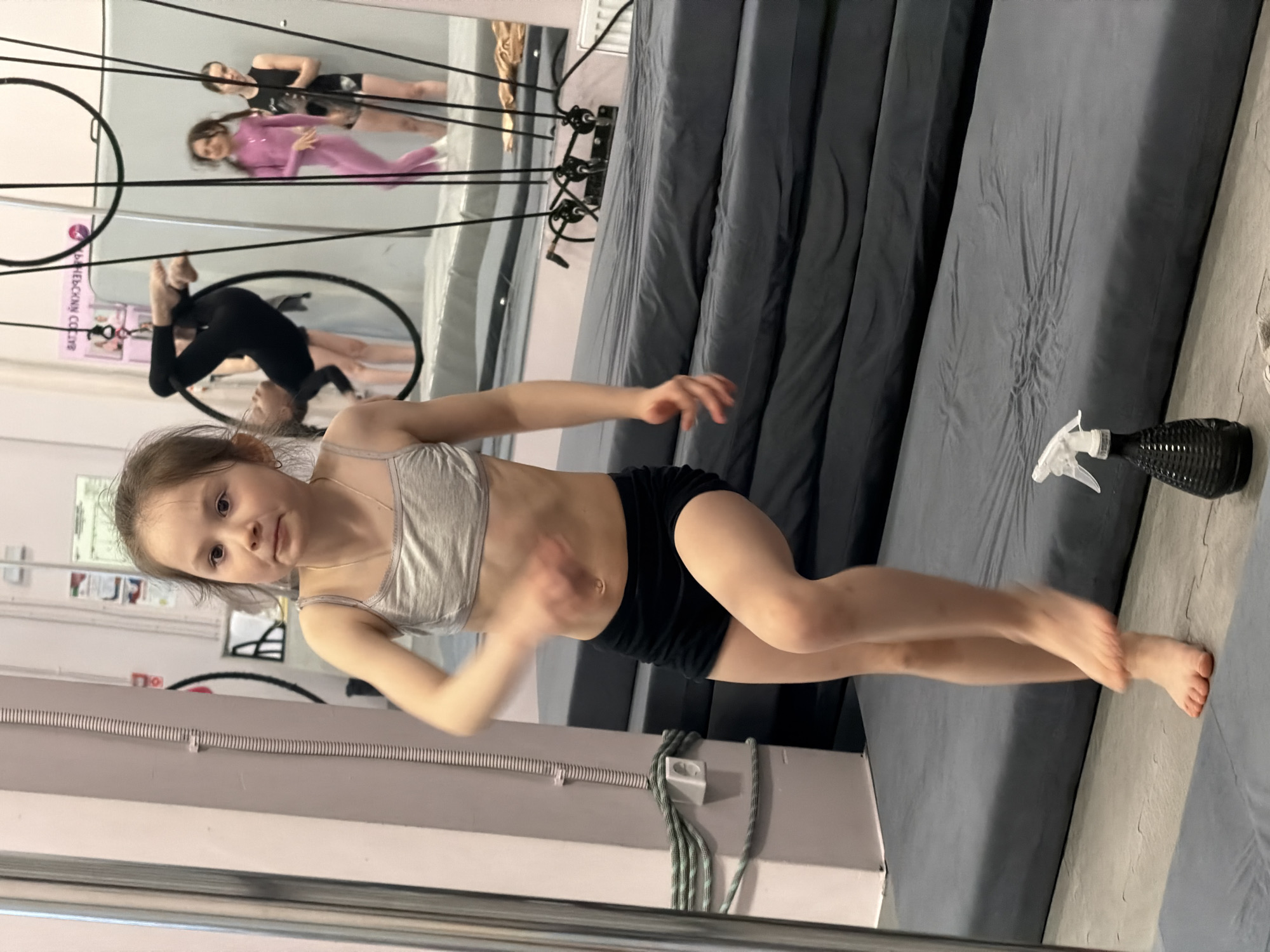 A girl in a gray sports bra and black shorts stands in front of a large mirror in a gymnastics studio, possibly located in Perm, Russia. Other individuals are visible in the mirror, some in aerial apparatuses. A gray padded surface is on the right side. A black spray bottle rests on the padded surface. Electrical wiring is visible along the wall on the left side.