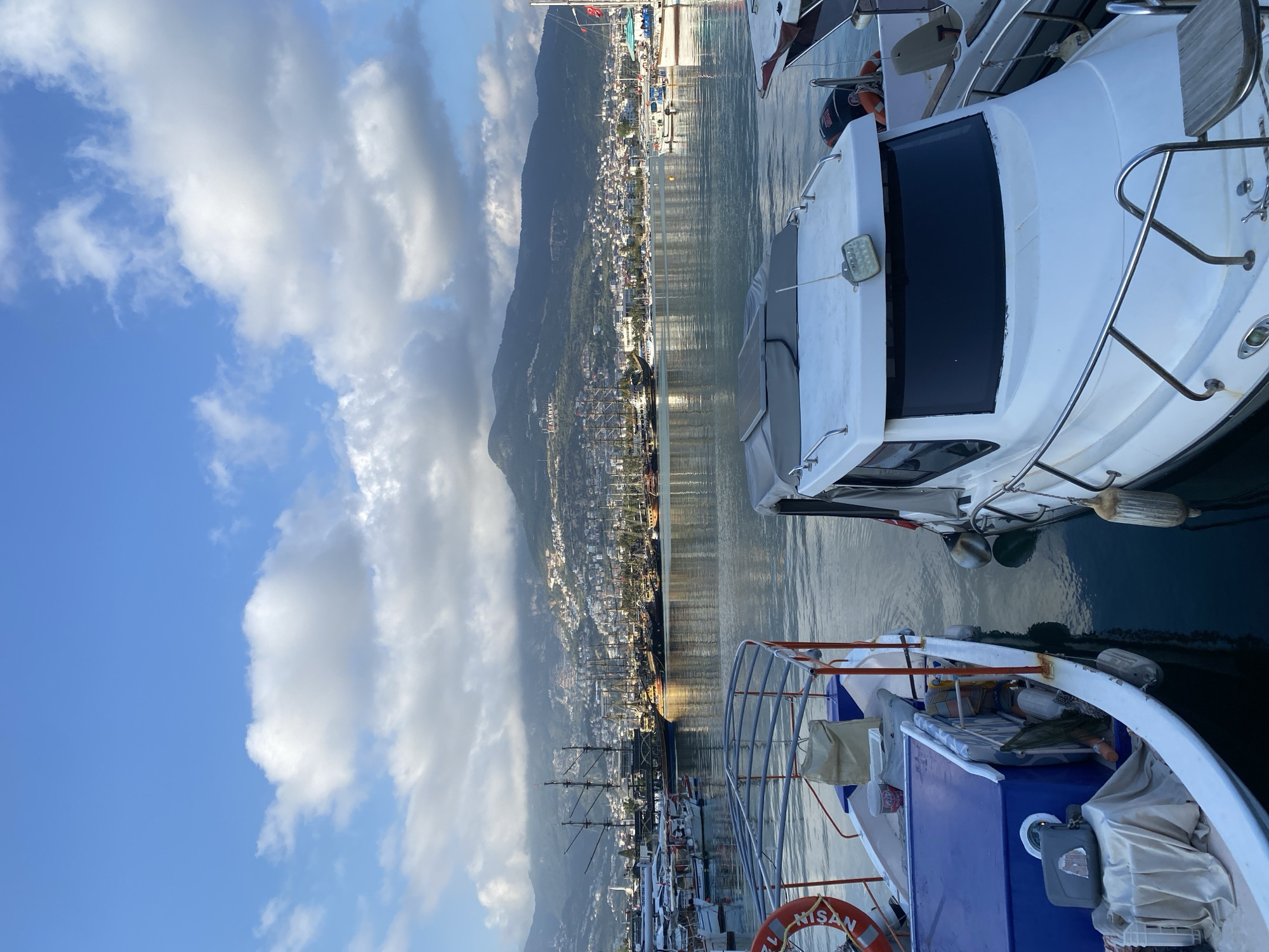 This image depicts a serene harbor scene in Alanya, Turkey, under a bright, partly cloudy sky. The perspective is from a dock, looking out across the marina towards a coastal city and mountains.
In the foreground, two boats are moored. To the immediate right, a white motor yacht displays its stern and part of its cabin, equipped with stainless steel railings. A luminous rectangular light fixture is visible on its side. To the left of this yacht, another boat, partially visible, features a white hull and a prominent blue deck structure. A red-orange lifebuoy with the text "NISAN" is attached to its side, and various covered items are stowed on its deck.
The calm waters of the marina fill the midground, reflecting the blue sky and the distant landscape. Several other vessels are visible further along the pier, including a distinctive large, dark-colored ship with multiple masts, resembling a pirate ship replica.
Dominating the background is the bustling coastal city of Alanya, sprawling along the base of a majestic mountain range. Numerous white and light-colored buildings are clustered, extending up the slopes. A partial view of the word "ALANYA" can be discerned on the lower part of the mountainside, confirming the location.
Above the city and mountains, a clear blue sky extends, adorned with scattered white and light grey clouds. The lighting suggests either early morning or late afternoon, with soft sunlight illuminating the scene and creating gentle reflections on the water. The overall impression is one of tranquility and natural beauty in a vibrant harbor setting.