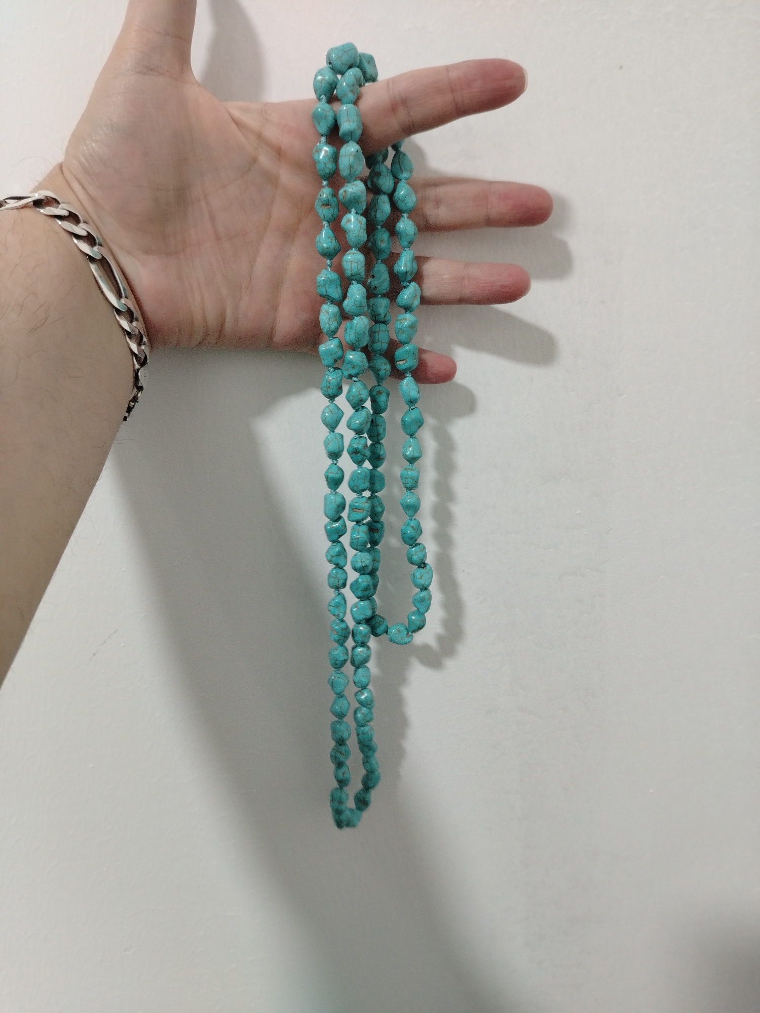 A person's hand is holding a long strand of turquoise-colored beads against a white wall. The hand is light-skinned and has a silver bracelet on the wrist. The beads are strung together and hang down in a triple strand. The background is a plain white wall. The location may be in Al Mas'oudi, United Arab Emirates.