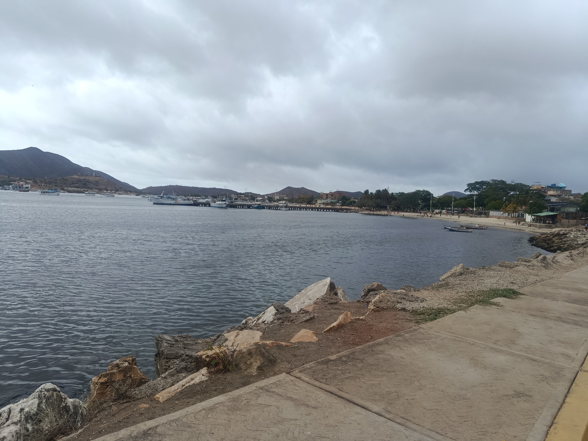 This image captures a wide view of the bay in Juan Griego, Venezuela, under an overcast sky. In the foreground, a concrete walkway with a patterned texture runs along the edge of the water. To the left, rough, rocky terrain with scattered boulders and dry vegetation meets the dark, rippling surface of the sea.
Along the shoreline in the mid-ground, a series of boats, including several white vessels and smaller fishing boats, are moored at piers and along the beach. The coastline features a sandy beach with scattered palm trees and vegetation. In the distance, low-lying hills and mountains form the backdrop, with buildings and structures visible along the shore and up the slopes. The water is a deep blue-gray, reflecting the muted light of the cloudy sky. A few people are visible on the beach, appearing to be walking or standing.