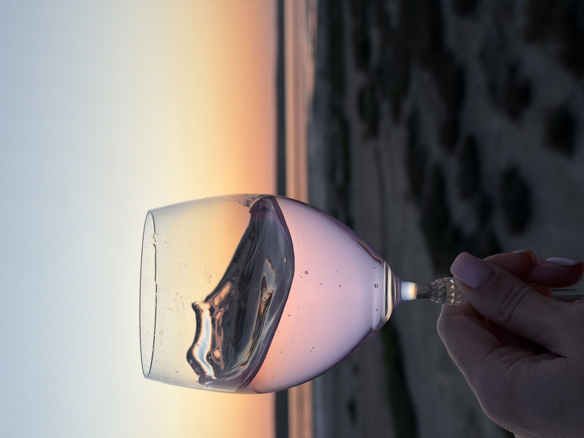 An outdoor shot captures a wine glass filled with a pale pink liquid, held by a hand with light pink fingernails. The liquid inside the glass is sloshing, creating a wave. The background shows a blurred landscape of dry vegetation and water under a twilight sky with hues of orange and blue. The scene appears to be set in the village of Korsuntsi, Ukraine, possibly near the Dniester River.
