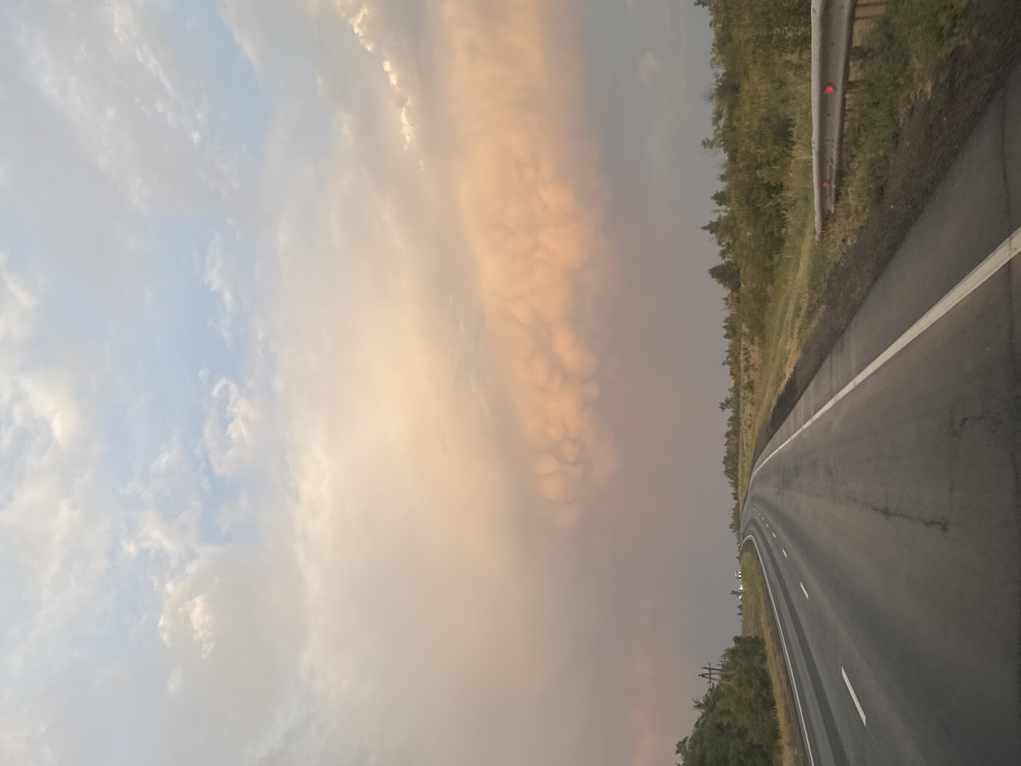 The image captures a view from a vehicle traveling on a paved road through a rural landscape, likely near Perevolotsky, Russia. The road, with white lane markings, curves gently into the distance. To the left, a grassy embankment with scattered trees lines the road. To the right, a metal guardrail with red reflectors is visible, bordering a similar grassy area with trees and bushes. The sky is filled with dramatic clouds, exhibiting a range of colors from light blue to pale orange and pink, suggesting either sunrise or sunset. The overall impression is one of a journey through a natural, open environment under an atmospheric sky.