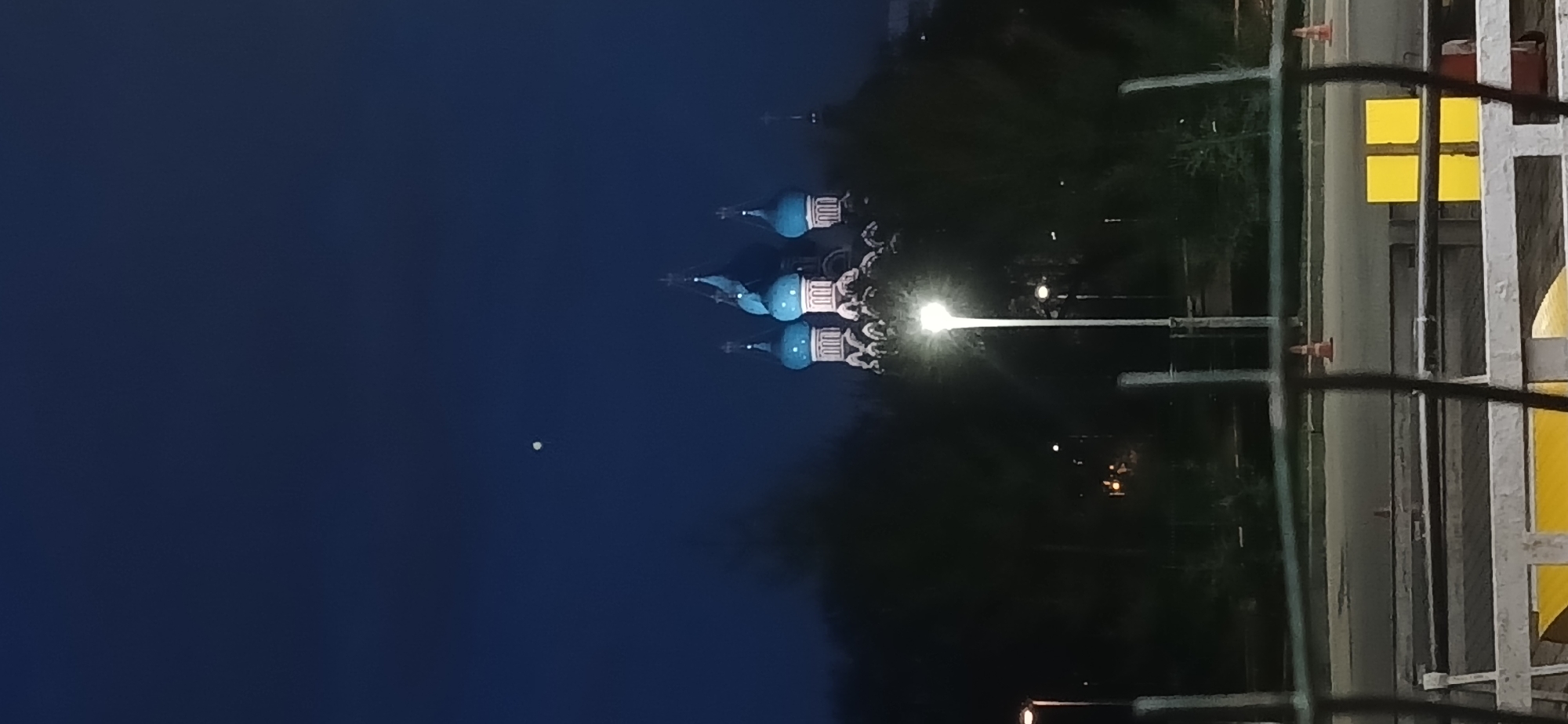 This is a nighttime shot of St. Basil's Cathedral in Moscow, Russia. The cathedral domes are blue with white stripes, and the sky is dark blue. A street lamp is visible, along with a portion of a metal fence and some yellow signage. The environment suggests an urban setting with park-like surroundings.
