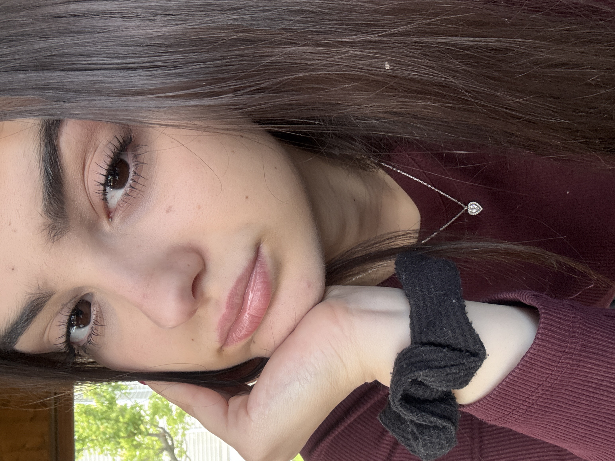 A young woman with brown hair is posing for a photograph. She is wearing a maroon sweater and a silver necklace with a heart pendant. Her left hand, adorned with a black scrunchie, supports her head. The background shows green foliage. The location of the image is unable to be determined.