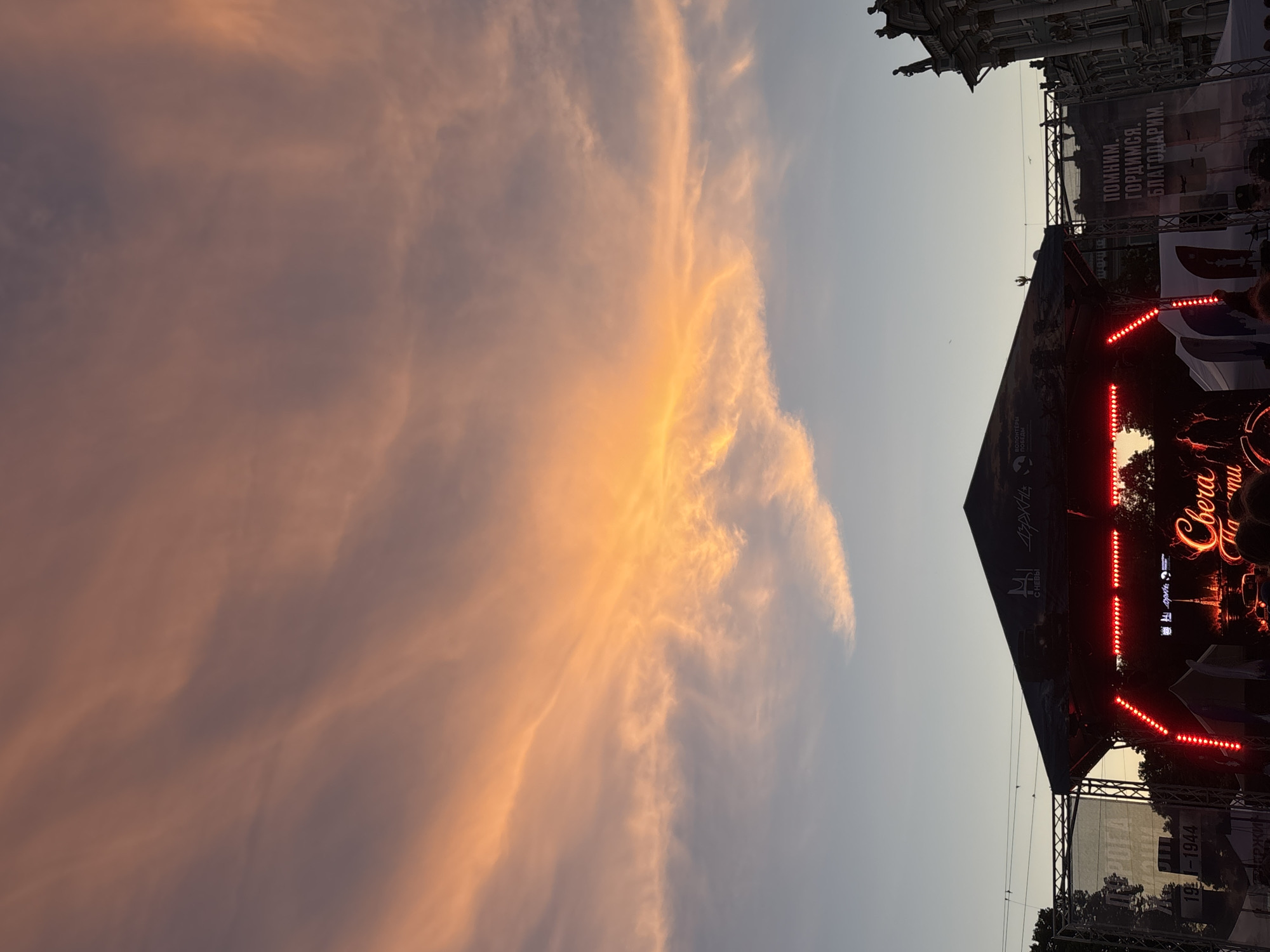 The image depicts a sunset over a street festival or concert in Saint Petersburg, Russia. The sky is dominated by dramatic clouds illuminated by the setting sun, casting a warm orange and pink glow. Below the sky, a temporary stage structure is set up, adorned with red LED lights. Behind the stage, banners with Russian text are visible, one of which reads "ПОМНИМ. ГОРДИМСЯ. БЛАГОДАРИМ." (We remember. We are proud. We are grateful.) and another referencing the years "1941-1944", likely related to World War II. A large building, potentially a historic structure like the Hermitage Museum due to its architectural style and placement, is visible on the right side of the frame, with statues on its roof. Silhouettes of people can be seen in the foreground, suggesting an audience attending the event. The overall atmosphere is one of a public gathering under a striking natural display.