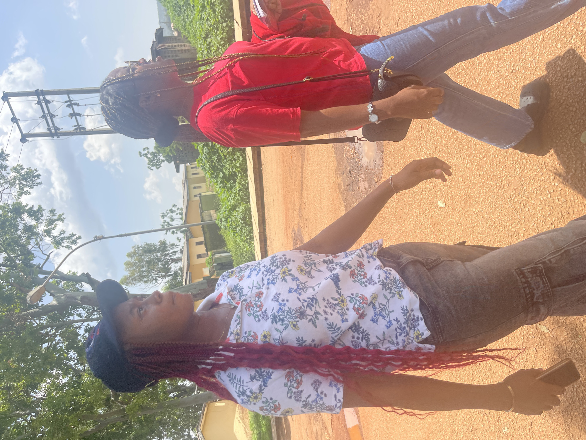 Two young women are captured in a vertical, outdoor photograph, likely taken in Rafin Par, Nigeria. The woman in the foreground, with long, burgundy braids and wearing a floral print shirt and dark jeans, is looking up and to her right. She has a black baseball cap on backwards, and her right arm is extended out and slightly bent. A silver bracelet is visible on her wrist, and she is holding a dark-colored smartphone in her right hand.
Behind her and slightly to the left, another young woman is also looking upwards and to her right. She has dark braids and is wearing a red t-shirt and blue jeans. A dark purse with a thin strap is slung over her shoulder.
The setting appears to be outdoors on a dusty, reddish-brown ground, possibly a path or road. In the background, there are green trees and some buildings, suggesting a semi-urban or rural environment. A utility pole with electrical wires is visible in the upper left portion of the image. The sky is bright with some clouds, indicating it is daytime. The lighting suggests it could be late morning or early afternoon, with the sun casting shadows on the ground. There is no visible text in the image. The overall mood of the image appears casual and relaxed.
