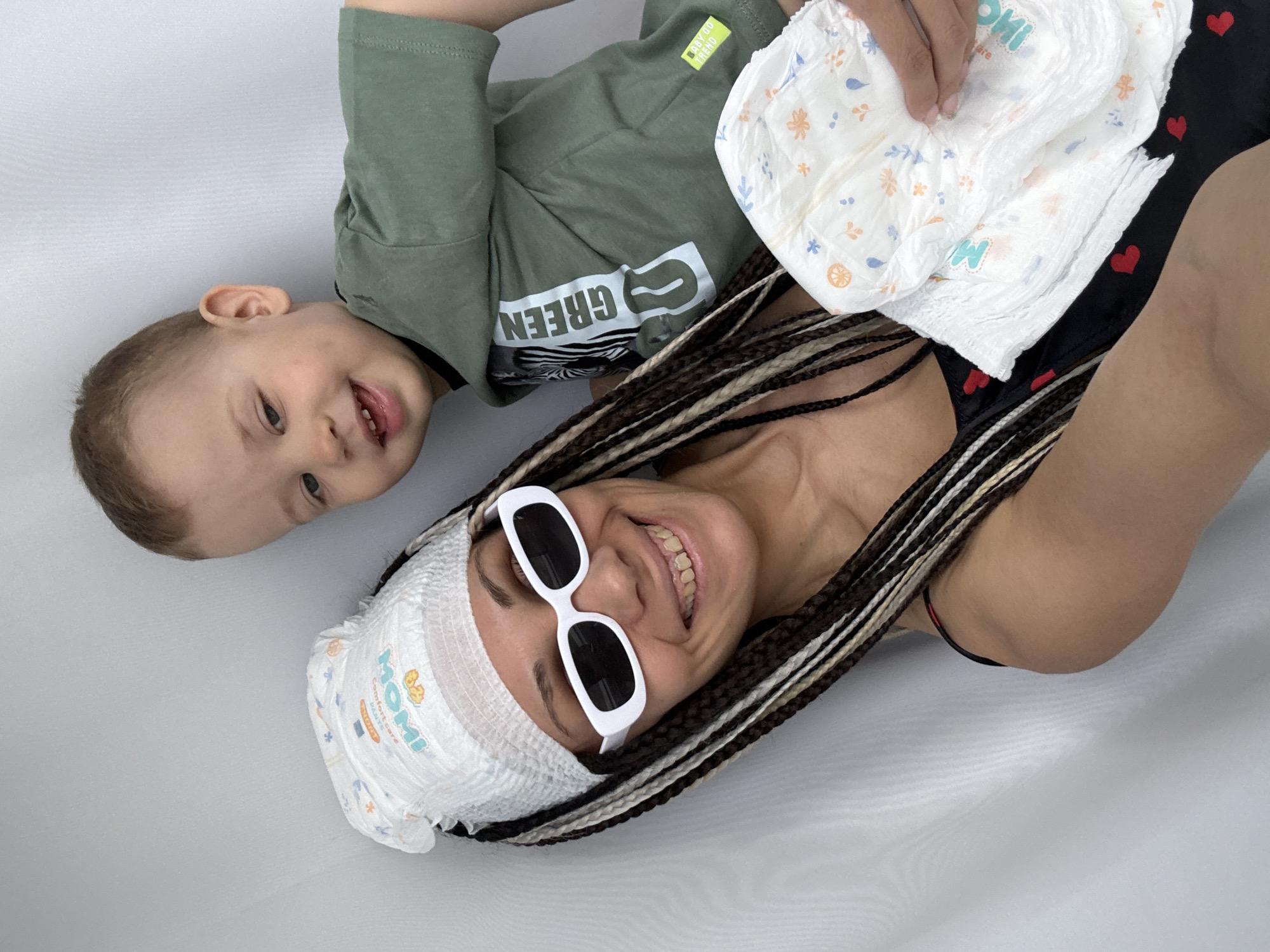 A woman wearing white sunglasses and a diaper wrapped around her head is lying on a white surface. She is smiling broadly. Next to her is a young boy in a green t-shirt, also smiling. The woman is holding a diaper in her right hand. The woman's hair is styled in long braids. The location is not specified in the image. The presence of children's diapers suggests a domestic or family-oriented setting.