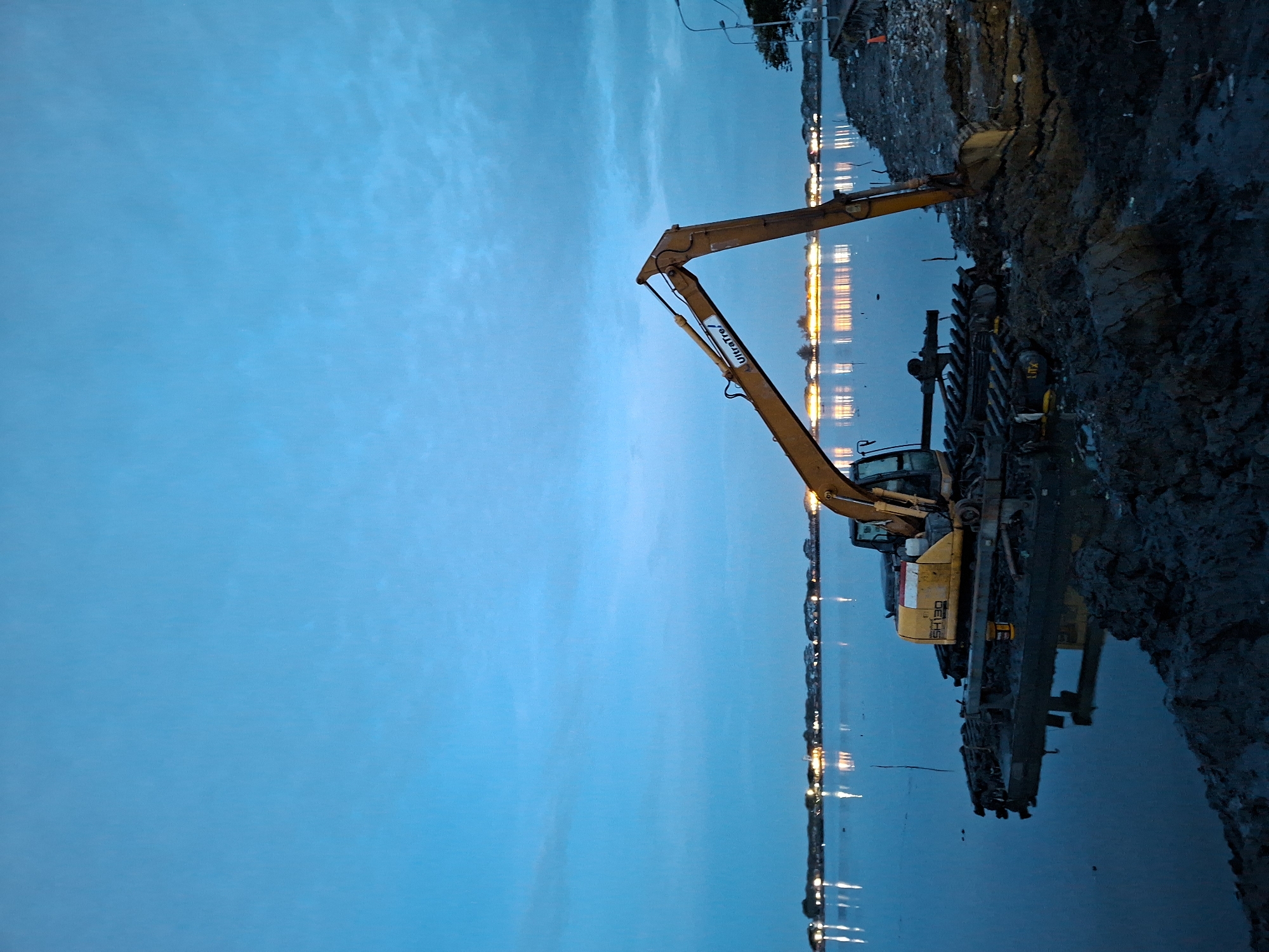 At dusk in Lhokseumawe, Indonesia, a yellow excavator arm stretches over water. The excavator sits on muddy land. Beyond the water, faint lights of the city twinkle on the horizon. The sky is predominantly blue.