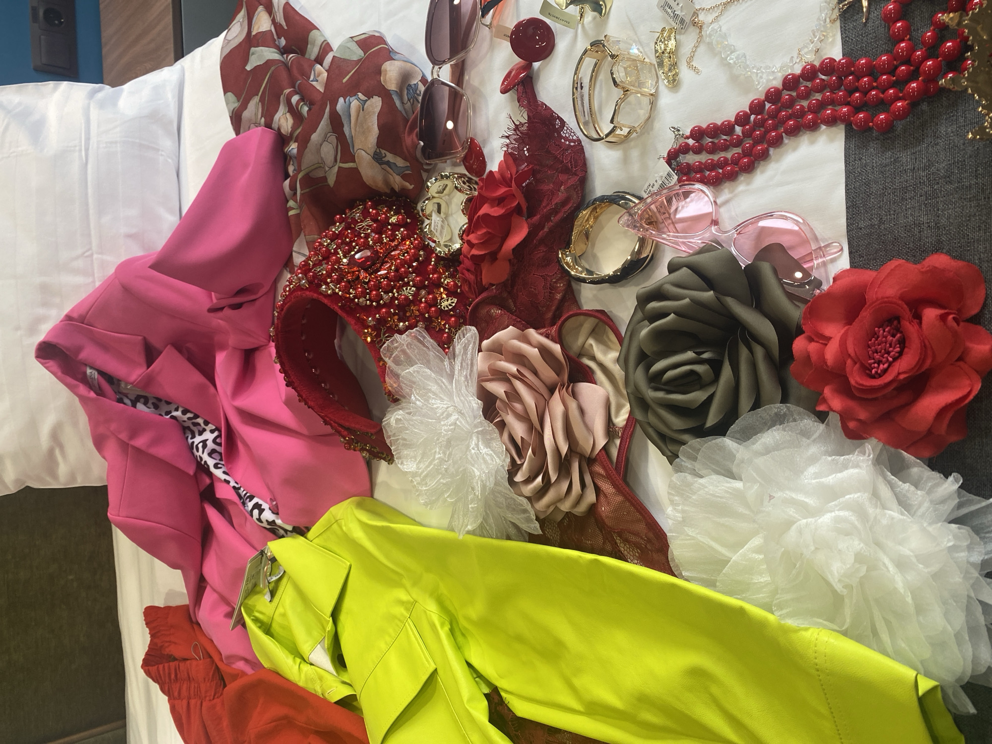 The image shows a collection of clothing and accessories scattered on a white bed. The clothing includes a bright pink blazer with a leopard-print lining, a neon yellow shirt, and a red garment. The accessories consist of sunglasses, jewelry, and various fabric flowers in shades of red, olive green, and white. A red beaded headband is also visible. The scene appears to be indoors, possibly in a hotel room or a personal residence. The setting is unknown and no city or location can be confirmed from the given image.