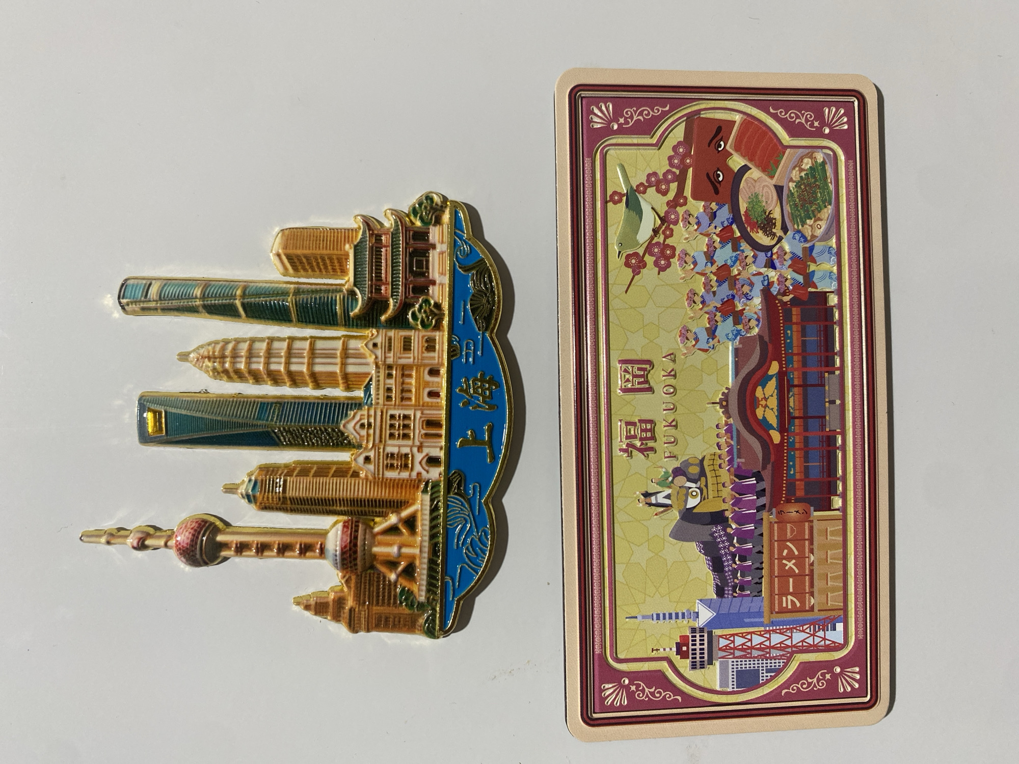 The image displays two souvenir items, likely magnets or cards, lying on a plain, light-colored surface.
On the left is a colorful, metallic magnet depicting the skyline of Shanghai, China. The magnet is oriented vertically but placed horizontally in the photograph. It features iconic skyscrapers such as the Oriental Pearl TV Tower, Shanghai Tower, Jin Mao Tower, and Shanghai World Financial Center, rendered in gold, brown, and green tones. A blue section, representing a river (likely the Huangpu River), flows past the buildings. The Chinese characters "上海" (Shanghai) are embossed on this blue section.
To the right is a vibrant, intricately illustrated postcard or souvenir card related to Fukuoka, Japan. This card is also oriented vertically but placed horizontally. It showcases various elements associated with Fukuoka, including a bird on a cherry blossom branch, a person in traditional Japanese attire, and multiple food items, prominently featuring ramen and other dishes. A lively scene with several small figures, possibly depicting a festival or street life, is also visible. Buildings and a cityscape form part of the background. The Japanese characters "福岡" and the English word "FUKUOKA" are clearly printed on the card. A sign on one of the buildings displays "ラーメン" (Ramen). The card has an ornate border with decorative patterns.
Both items are static and there is no activity taking place in the scene itself, other than being photographed. The items are souvenirs representing specific cultural and architectural landmarks of Shanghai, China, and Fukuoka, Japan. The photo was taken in an unspecified location, though the user provided "Verkhnevesyoloye, Russia" as a context for where the photo was taken, not what the items represent.