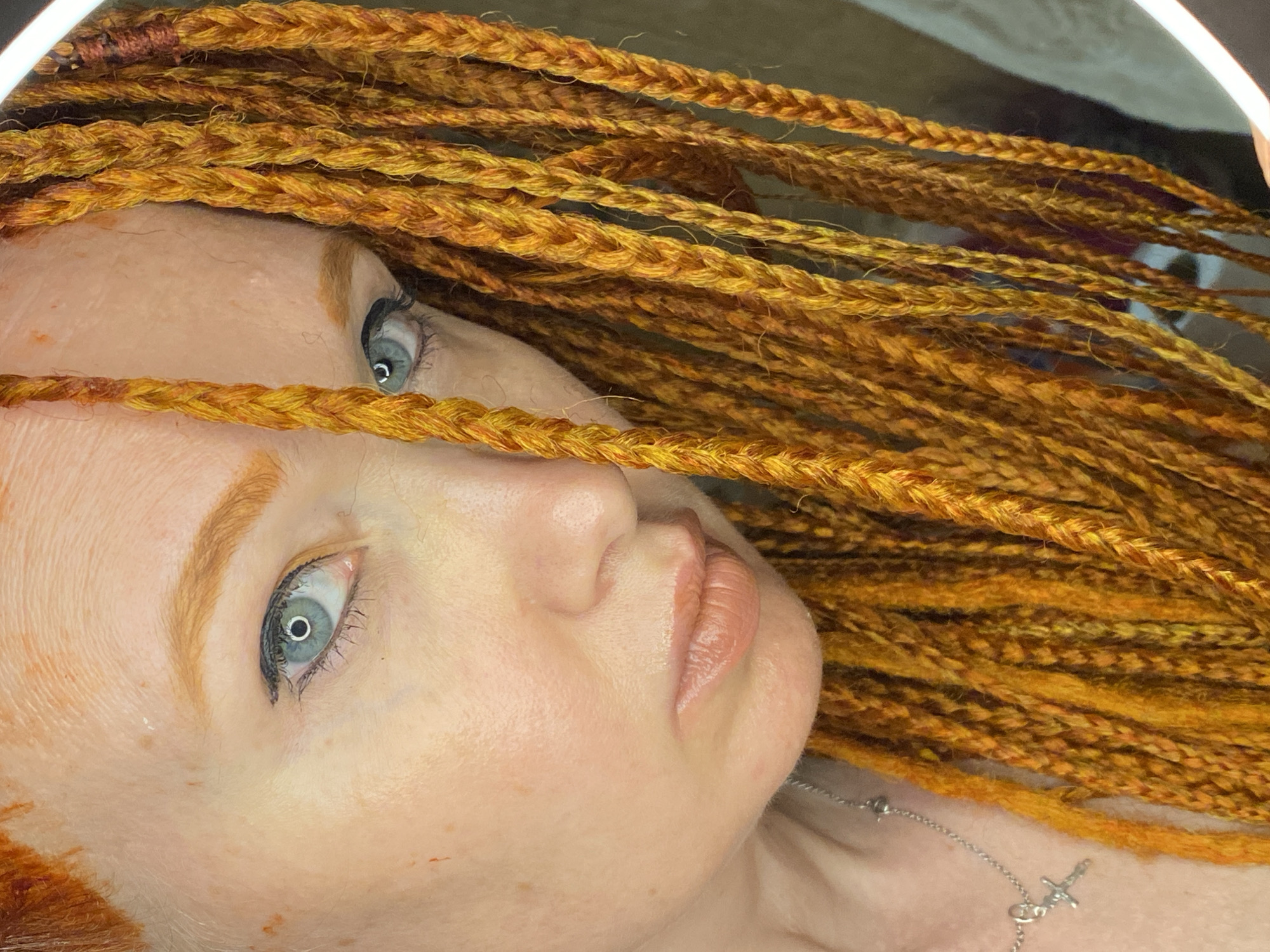 The image shows a close-up of a woman's face, with her head turned to the side. She has pale skin with freckles, light blue eyes with black eyeliner, and light brown eyebrows. Her hair is a vibrant orange, styled in numerous thin braids that partially cover her face. A silver necklace with a small cross pendant is visible around her neck. The background is out of focus and appears to be indoors, with a hint of a white ring light reflecting on her forehead and to the right of the image. There is no indication of the city of Sochi, Russia, or any other specific location.