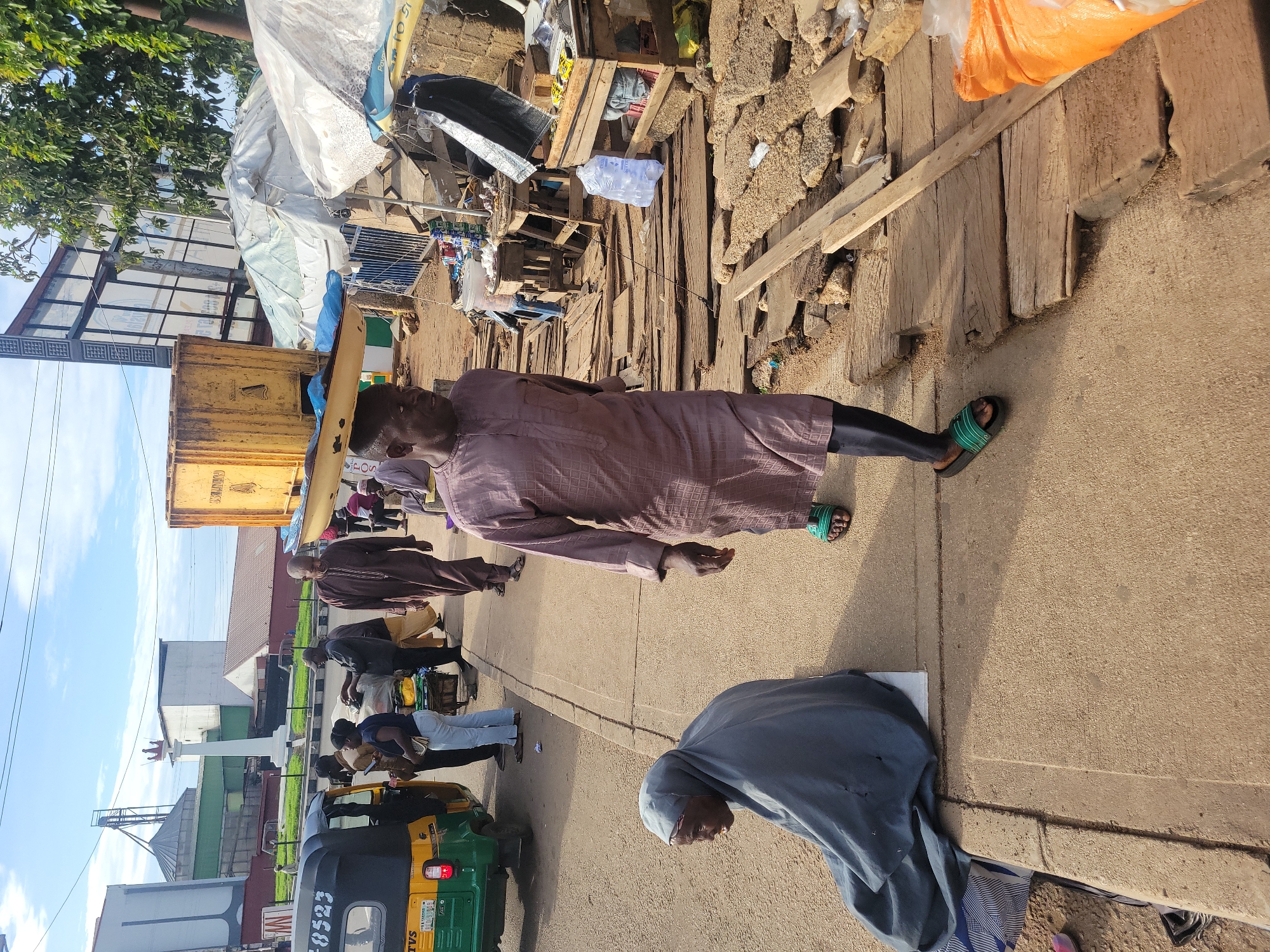 This image captures a daytime urban street scene in Jos, Nigeria, likely near the Jos Museum Roundabout, identifiable by the distinctive white monument topped with a crown in the background.
In the immediate foreground, a man, dressed in a patterned purple tunic and dark trousers with green and black sandals, walks rightward on the edge of the asphalt road, balancing a flat, light-colored object on his head. To his left, on a concrete sidewalk, an older woman cloaked in a grey garment and head covering is seated directly on the ground, facing left.
In the mid-ground on the left, a green and yellow TVS King auto-rickshaw, bearing the license plate "PLATEAU HNG 8523," is parked or stopped. A small group of individuals, including a woman in light blue jeans and a dark top, stands on the sidewalk near the auto-rickshaw.
The mid-ground on the right features a makeshift market area with several stalls constructed from wooden frames and covered with white and blue tarpaulins or fabrics. Various goods, including packaged items, a clear plastic bag of water, and an orange sack, are visible on or around wooden tables. The ground in this market section is unpaved and uneven, composed of loose dirt, wooden planks, and stones. Several people are present within and around these market stalls, some appearing to be vendors or customers.
In the background, behind the monument, large industrial-style buildings, possibly silos, are visible under a partly cloudy blue sky. A large billboard structure is partially discernible on the far right. Overhead power lines traverse the upper portion of the scene. The lighting indicates bright daylight conditions.