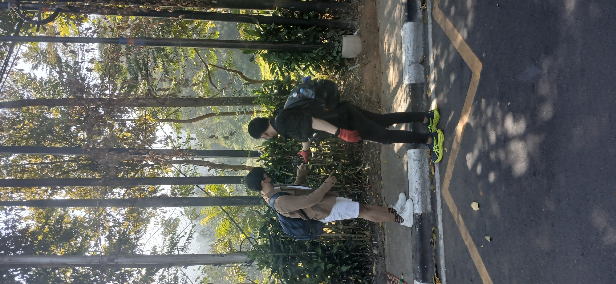 Two individuals are standing on a paved sidewalk adjacent to a road, with a line of trees and lush green foliage in the background. The individual on the left is wearing a light brown jacket, white shorts, and white sneakers with dark striped socks. They have a blue backpack on their back and are holding a smartphone. The individual on the right is wearing a black long-sleeved shirt, black pants, and bright yellow and black sneakers. They are also wearing red gloves and have a black backpack with a water bottle attached. They are facing the individual on the left and appear to be holding a red object, possibly a phone or a camera, out towards them. The road has yellow lane markings. The environment appears to be outdoors, with sunlight filtering through the trees, casting shadows on the ground. The context suggests this scene is located in Cisarua, Indonesia, likely an area with dense vegetation and roads.