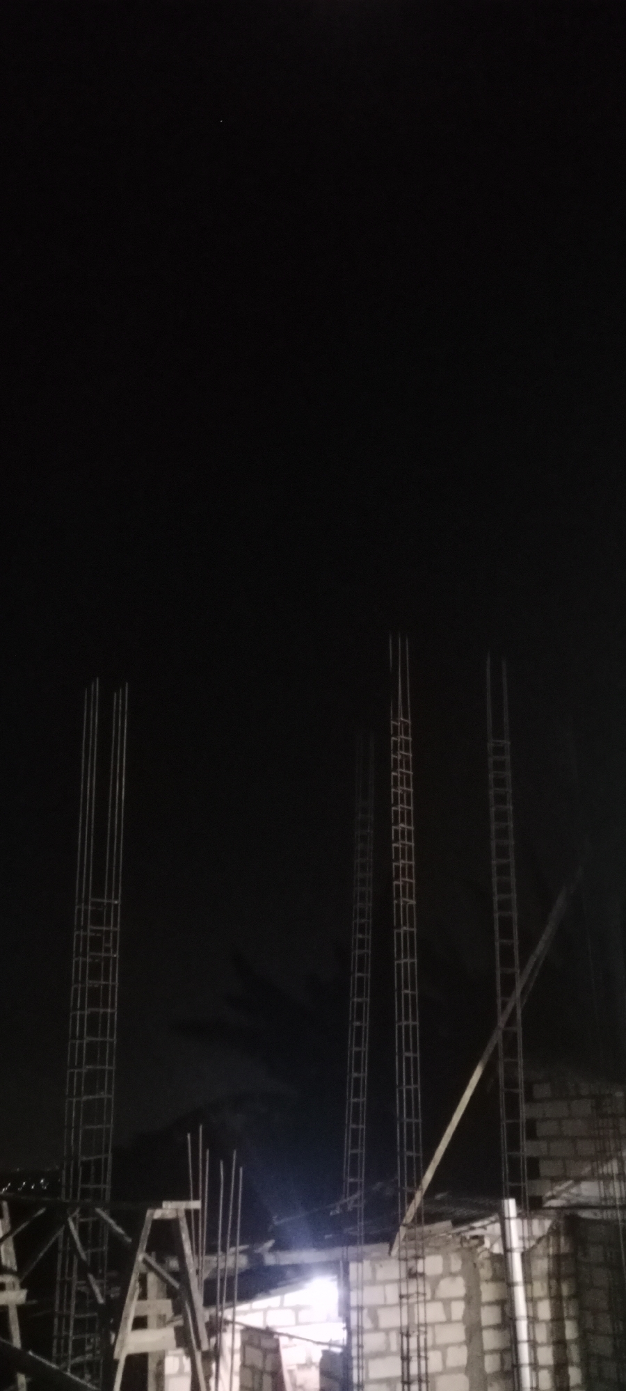 A construction site is visible at night in Soko, Indonesia. The scene is dark with some artificial lighting creating highlights and shadows. Several vertical rebar structures, appearing as columns, are prominent in the foreground. In the background, brick walls of a partially constructed building are visible. The sky is dark, with a faint cloud formation. There are no people visible in the image. The overall impression is of an ongoing construction project at night.