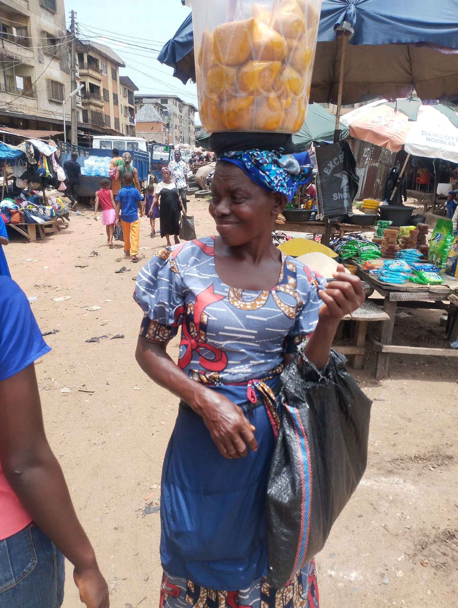 In the Fegge district of Nigeria, a woman balances a transparent container filled with yellow fruits on her head. She wears a blue headscarf, a patterned blue top, and a blue skirt, carrying a black bag. The environment is a bustling marketplace with stalls, buildings in the background, and people passing by. A Dangote Noodles umbrella and various goods are visible on the stalls.