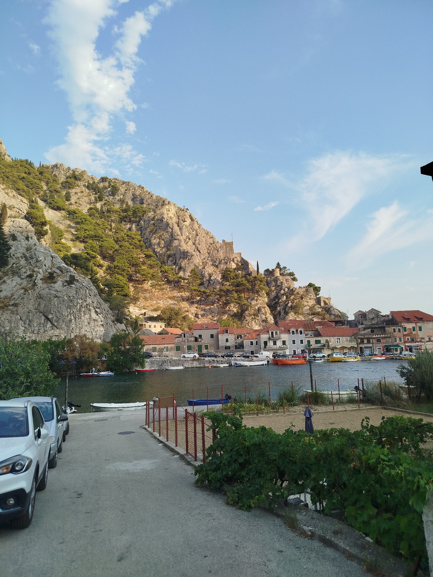 The image depicts a scenic view of Omiš, Croatia, a coastal town situated at the mouth of the Cetina River, nestled at the foot of steep, rocky cliffs. The foreground shows a paved road with several cars parked along its left side, including a white SUV. To the right, a red-railed fence separates the road from a gravel area with lush green foliage in the immediate foreground.
Beyond the gravel area, the Cetina River flows, lined with a waterfront promenade. Numerous boats of various sizes and colors are moored on the river, including bright orange and yellow vessels, as well as smaller white dinghies. On the opposite bank of the river, a charming cluster of buildings with red-tiled roofs forms the town's picturesque skyline. These structures range from multi-story houses to smaller, more traditional dwellings.
Towering above the town are the imposing, rugged cliffs characteristic of Omiš. The slopes are partially covered with dense green vegetation, including pine trees. Atop the highest cliff, the remains of a medieval fortress, likely the Mirabela Fortress, are visible, adding a historical dimension to the landscape. The sky above is a clear blue, dotted with scattered white clouds, suggesting fair weather. The overall impression is one of a tranquil, picturesque coastal town with a rich history and a dramatic natural setting.