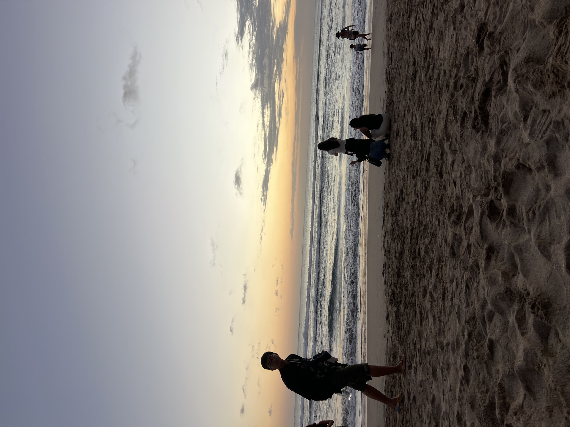 The image depicts a beach scene in Kuta, Indonesia, during sunset. The sky displays a gradient of colors from light blue at the top to warm orange and yellow hues near the horizon. Several people are present on the sandy beach. In the foreground, a man walks towards the left side of the frame, wearing a dark jacket and shorts. Further back, closer to the water, a group of individuals is gathered. Three women are crouched down, possibly taking photos or socializing. Another woman stands behind them, facing the ocean. To the right, a child runs near the water's edge, with another person in the background walking along the shore. The ocean has moderate waves, and there are some small boats visible on the water in the distance. The sand is a rich brown color, with visible footprints and textures.