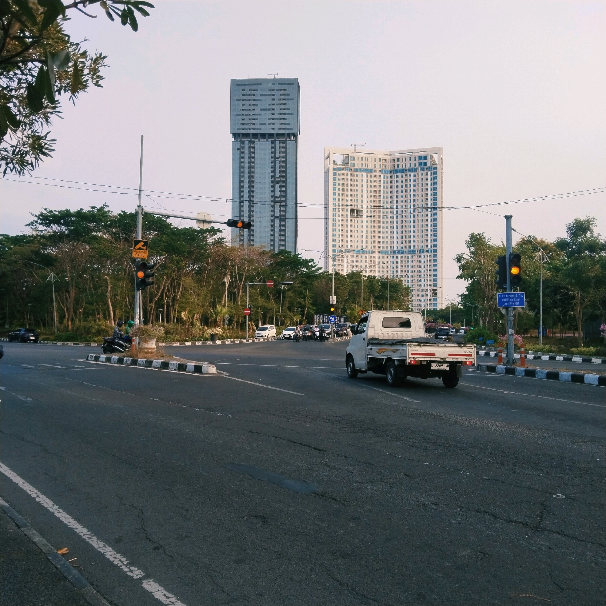 The image depicts a street scene in Surabaya, Indonesia, likely at an intersection. In the background, two prominent high-rise buildings stand against a clear sky. One building is a sleek, modern skyscraper with a distinctive angular top, and the other is a wider, curved building with numerous windows. In the middle ground, a dense line of trees borders the road, with some bare branches visible.
The foreground shows a paved road with white lane markings and some visible cracks and wear. A white pickup truck is driving on the right side of the road, moving away from the viewer. To the left of the truck, traffic lights are visible, with the yellow light illuminated. A traffic sign with "AREA DILARANG CCTV" (CCTV PROHIBITED AREA) is mounted on a pole next to the traffic light.
Further back, several motorcycles and a few cars are present, suggesting moderate traffic. Two people are on a motorcycle, and another is on a separate motorcycle. The road is bordered by a concrete divider with black and white striped markings, and a small planter with foliage is situated on this divider.
The overall scene suggests an urban environment during daylight, possibly late afternoon due to the lighting and shadow conditions. The image has a slightly muted color palette.