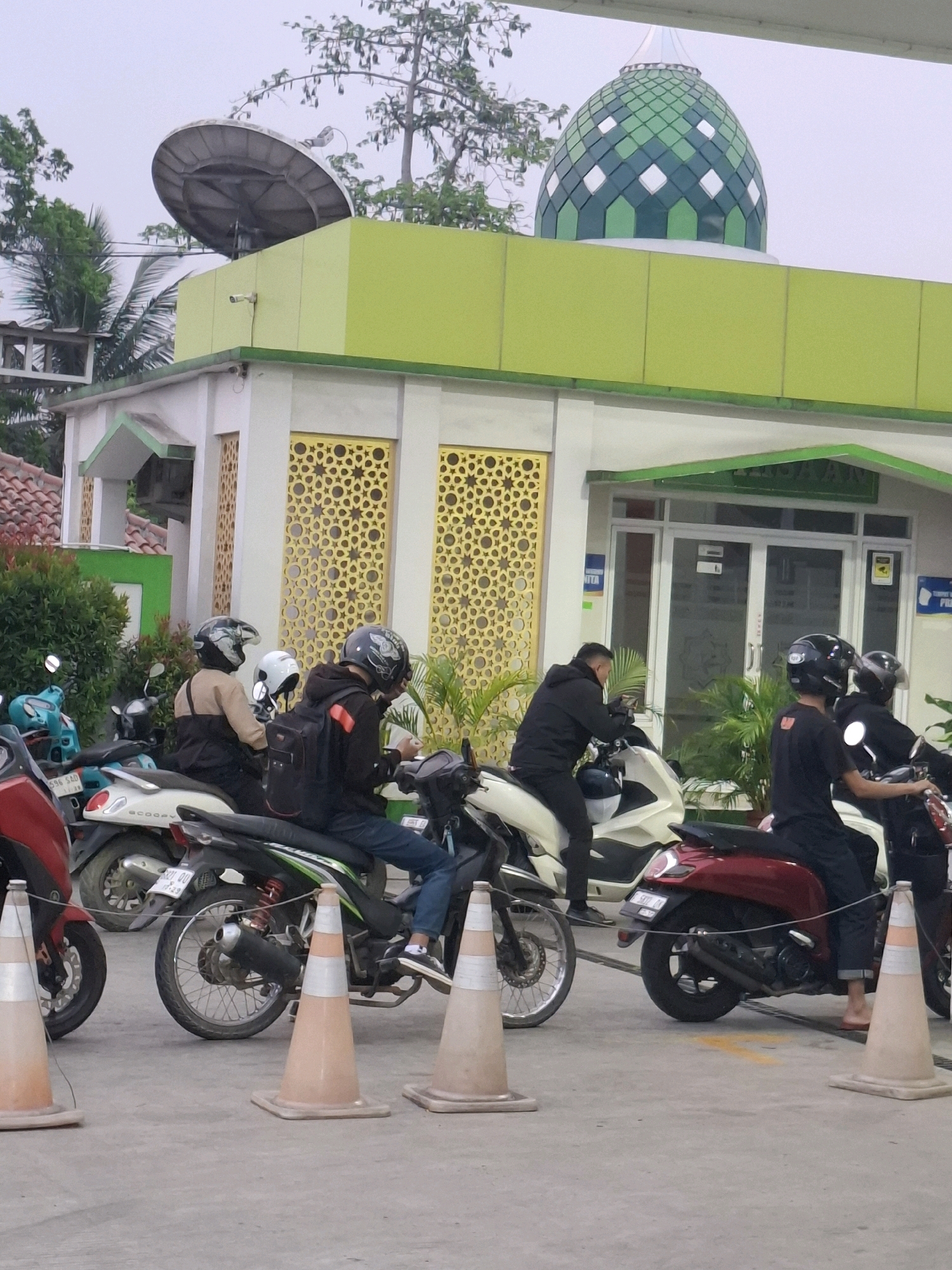 The image captures an outdoor scene, likely a gas station, in Sukabumi, Indonesia. A building with a lime green trim and white facade is visible, featuring decorative gold patterns on its walls and a green domed roof. Several people are present, most of whom are on or around motorcycles. One person in the foreground is seated on a red scooter, facing left, with their feet bare. To their left, a person on a white scooter is facing forward. In the middle ground, two individuals are on a white scooter, one driving and the other seated behind. To the right, another person is on a black scooter, also facing forward. The ground is paved, and traffic cones are placed around the area. In the background, a satellite dish is mounted on the roof of a building, and trees are present. The sky is overcast.