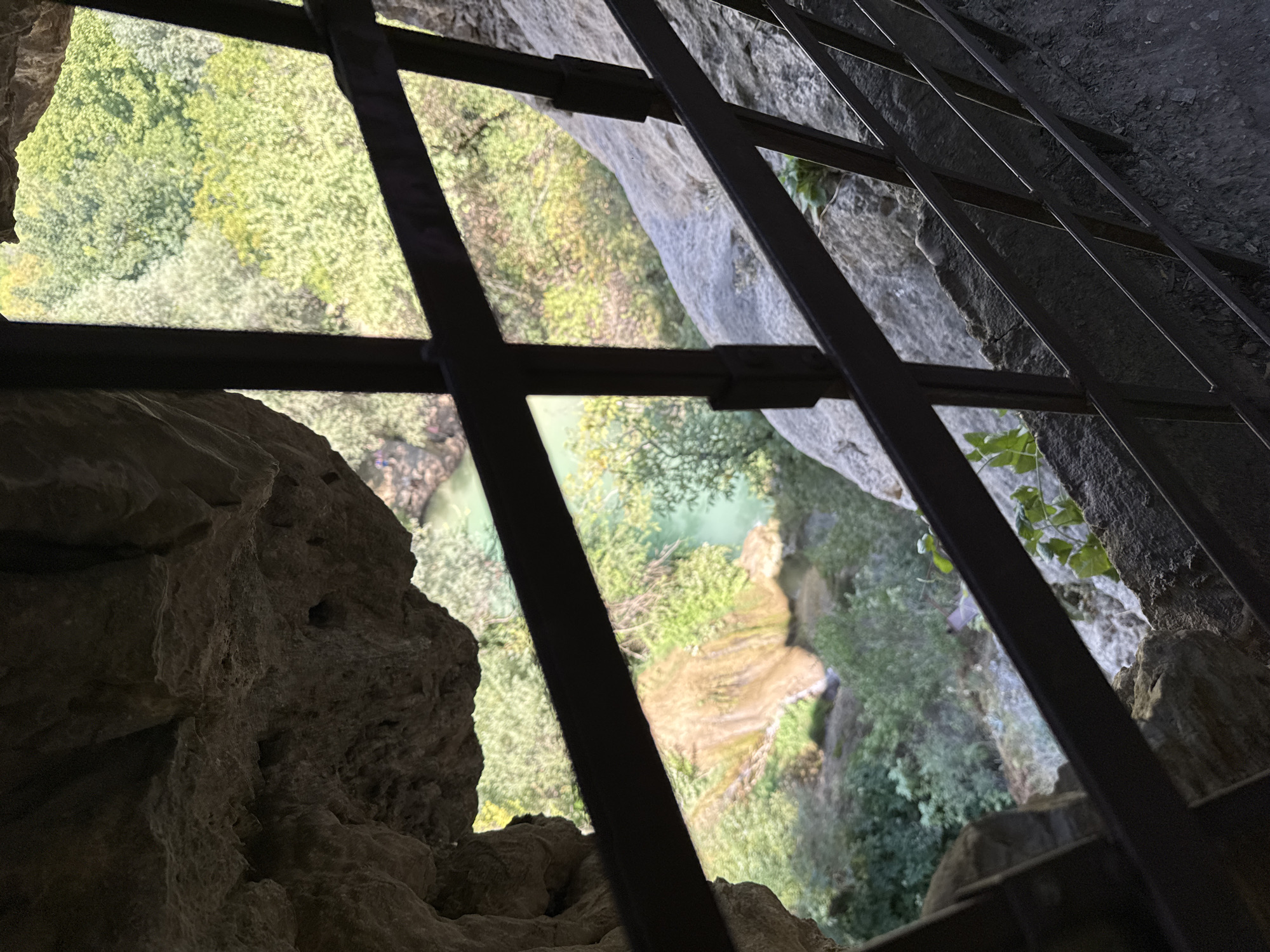 The image shows a view through a metal railing and a rock overhang, looking out at a waterfall and lush green vegetation. The perspective is from a cave-like opening. The railing consists of horizontal bars and vertical supports. The rock overhang is rough and textured, with shadows suggesting depth. Below and in the distance, a waterfall cascades down a rocky slope into a turquoise body of water. The surrounding area is densely forested with trees showing various shades of green. The scene appears to be a natural landscape. This viewpoint is characteristic of the "Cave at the Waterfall of Hotnitsa" located near the village of Hotnitsa in Bulgaria.