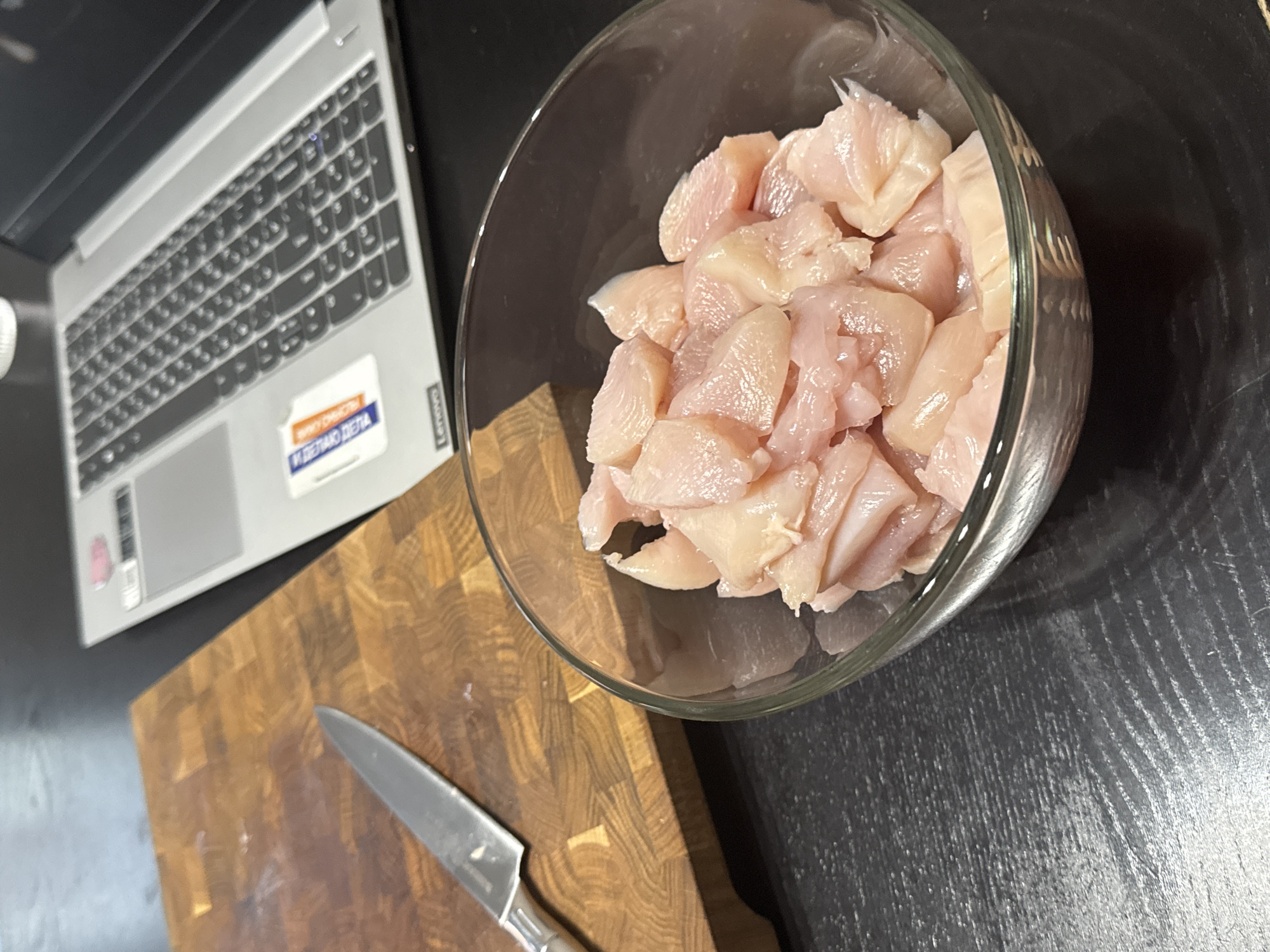 The image shows a kitchen scene with a laptop, a bowl of diced chicken, a cutting board, and a knife. The laptop is a Lenovo, indicated by a sticker. The cutting board is wooden, and the knife is a stainless steel chef's knife. The chicken is in a clear glass bowl. The background appears to be a black kitchen countertop. There is a sticker on the laptop with the words "МЫ ДЕЛАЕМ ДЕЛА" which translates to "WE DO BUSINESS." This sticker suggests the scene might be located in Russia. The Russian writing on the sticker would suggest a location in Russia, possibly the city of Perm.