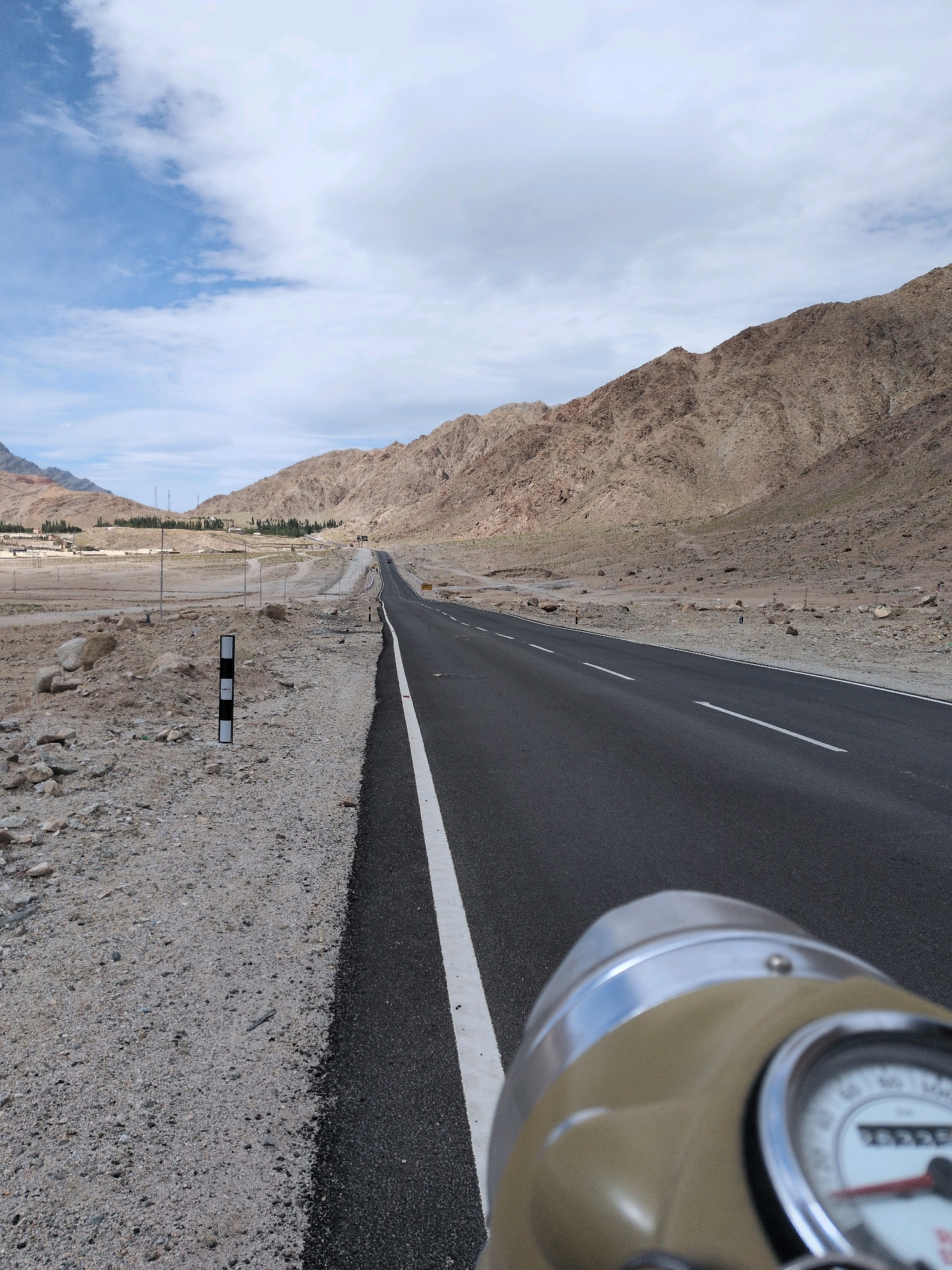 The image depicts a desolate, arid landscape in Upshi, India, characterized by rugged, brown mountains that dominate the background. A paved asphalt road with a white dividing line stretches into the distance, winding through the valley. The immediate foreground shows a rocky, gravelly terrain. On the left side of the road, a black and white striped marker post is visible. In the far distance, a cluster of buildings and some sparse greenery suggest a small settlement. The sky is a mix of blue and white clouds. The shot appears to be taken from the perspective of someone riding a motorcycle, as the handlebar and a portion of the speedometer are visible in the lower right corner of the frame.