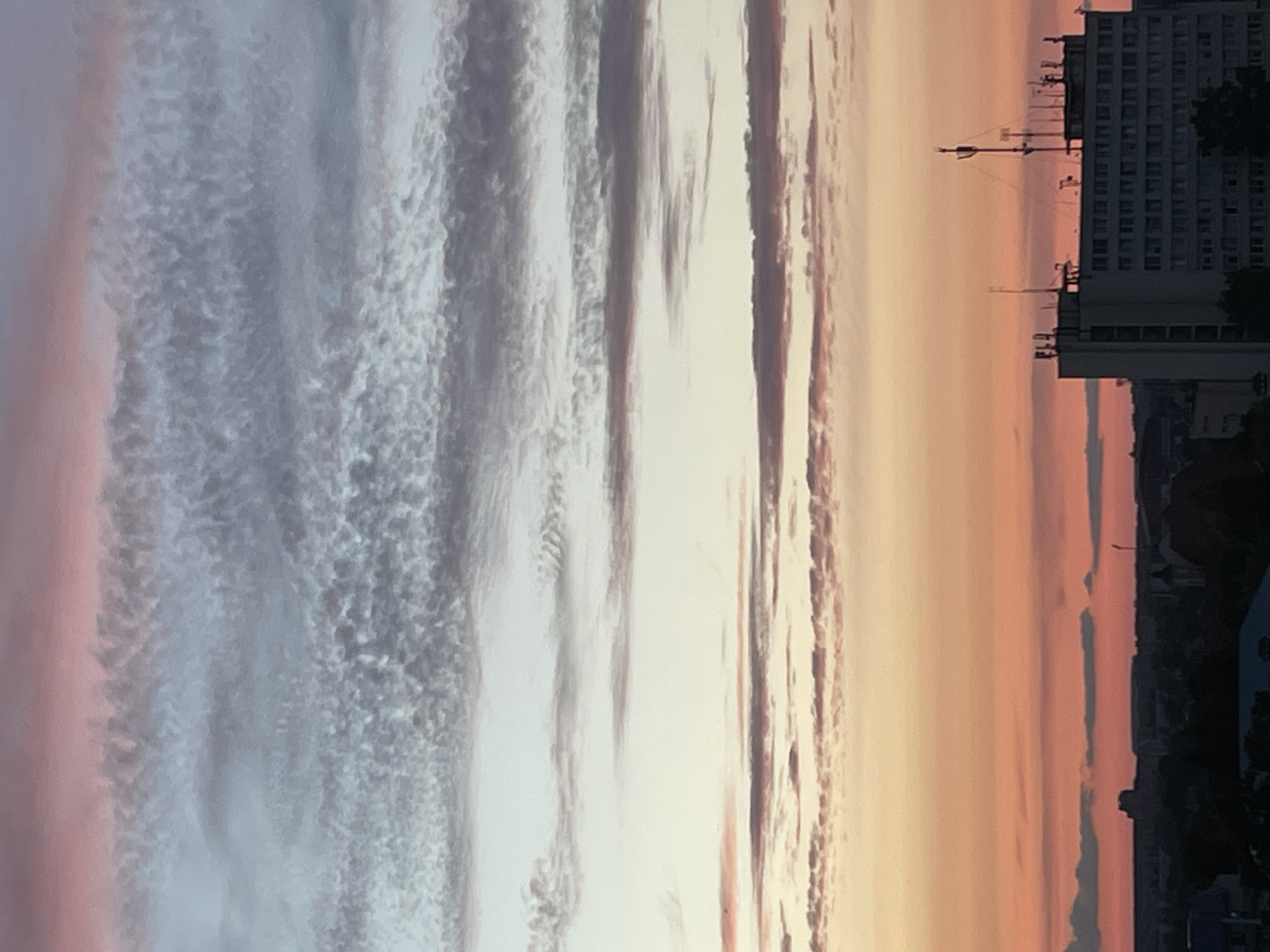 The image depicts a sky with sunset colors over a city skyline, specifically in Hrodna, Belarus. The sky is filled with multiple layers of clouds, exhibiting hues of pink, orange, yellow, and blue. The upper portion of the sky features a dense layer of small, textured clouds in shades of grey and pink, transitioning to lighter blues and yellows lower down. A distinct band of orange and pink clouds sits just above the horizon. In the foreground, a multi-story building with many windows is visible, topped with various antennas and communication equipment. The silhouette of other buildings and structures can be seen on the horizon, along with dark trees. The lighting suggests it is either sunrise or sunset. There are no people visible in the image.