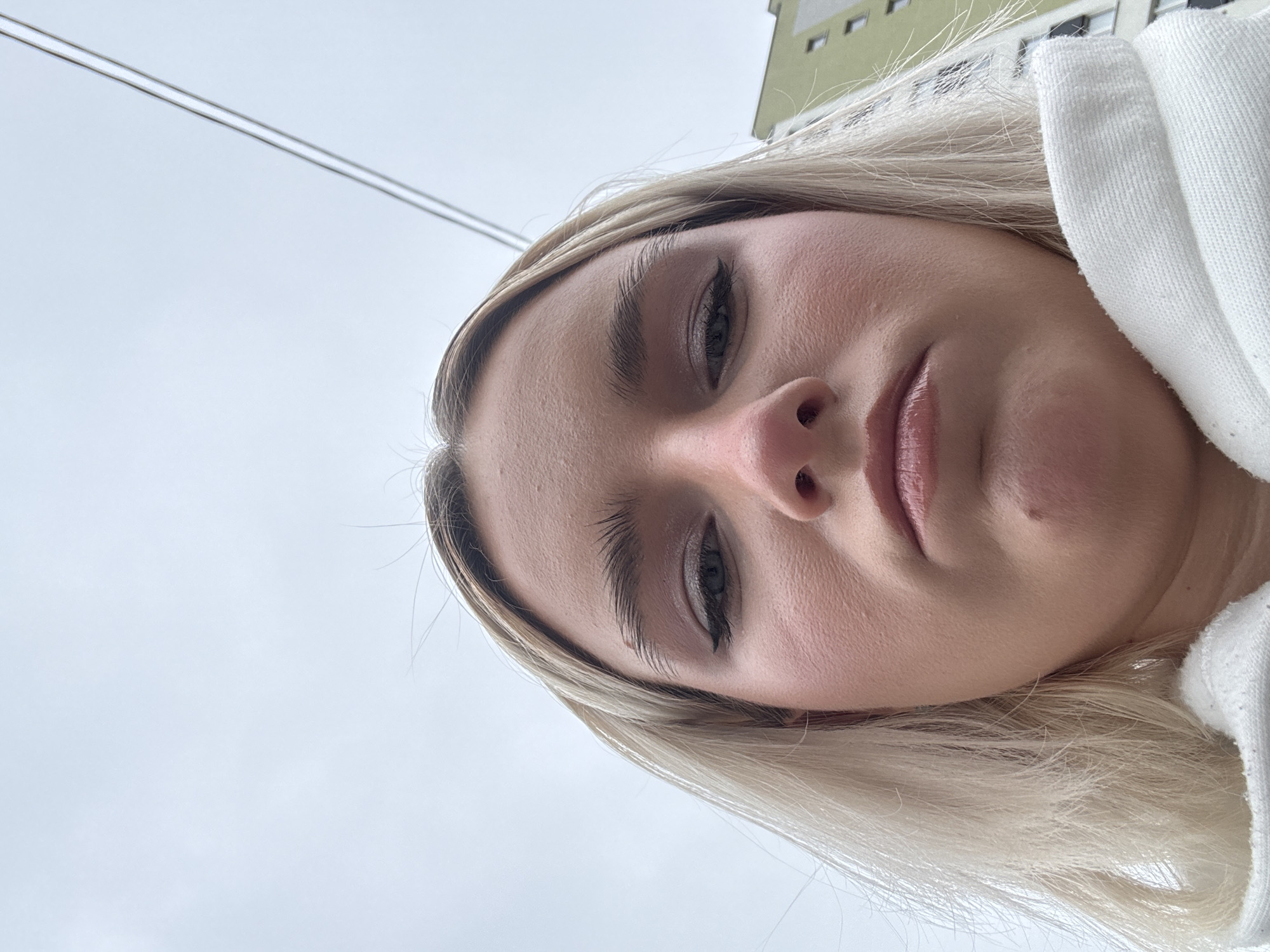 The image shows a close-up, low-angle selfie of a woman with blonde hair. Her face fills the majority of the frame, with her eyes directed downwards. She appears to be wearing eyeliner and lipstick. In the background, a light gray sky is visible. At the top edge of the image, there is a portion of a building with green siding. Above her head, parallel lines likely indicate power lines. A part of a white garment is visible on the right side. The image was taken in Unknown, Russia.