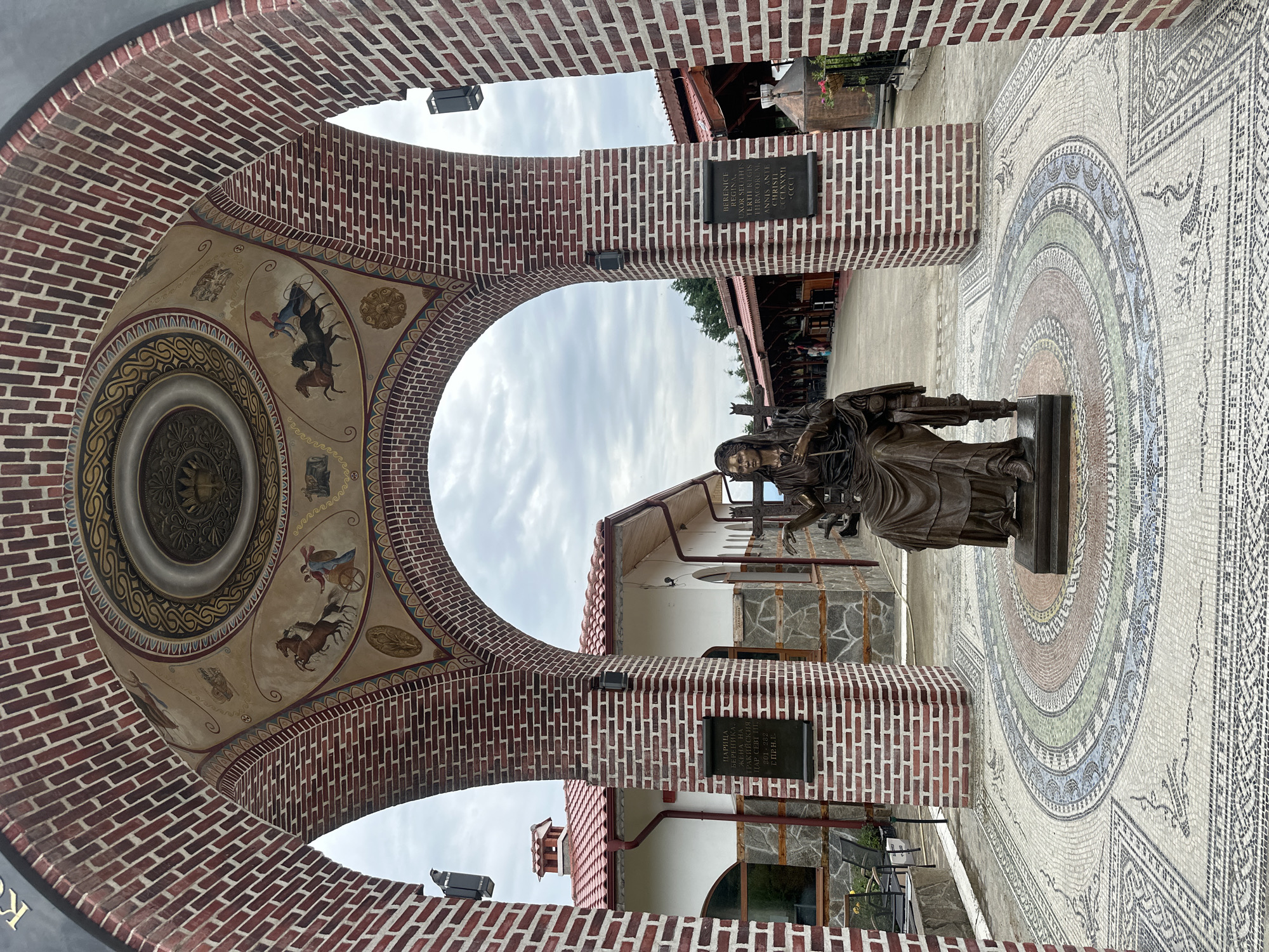 This media file shows a view through a brick archway into an outdoor courtyard. The archway itself is decorated with a fresco on its ceiling, depicting scenes of chariots pulled by horses and other figures. The courtyard floor features a colorful mosaic with geometric patterns and a large circular design.

In the center of the courtyard stands a bronze statue of a woman, possibly a historical figure, holding a book or scroll and a cross. The statue is placed on a pedestal, and behind it, a building with a red-tiled roof is visible. The building's facade is a mix of white walls and stone accents.

On one of the brick pillars supporting the archway, there is a black plaque with text in Cyrillic and Latin script. The text appears to be a historical inscription, possibly related to the statue or the location. The sky is overcast, suggesting a cloudy day. The overall scene evokes a sense of historical significance and artistic appreciation, likely a cultural or religious site. The location is identified as Skobelevo, Bulgaria.