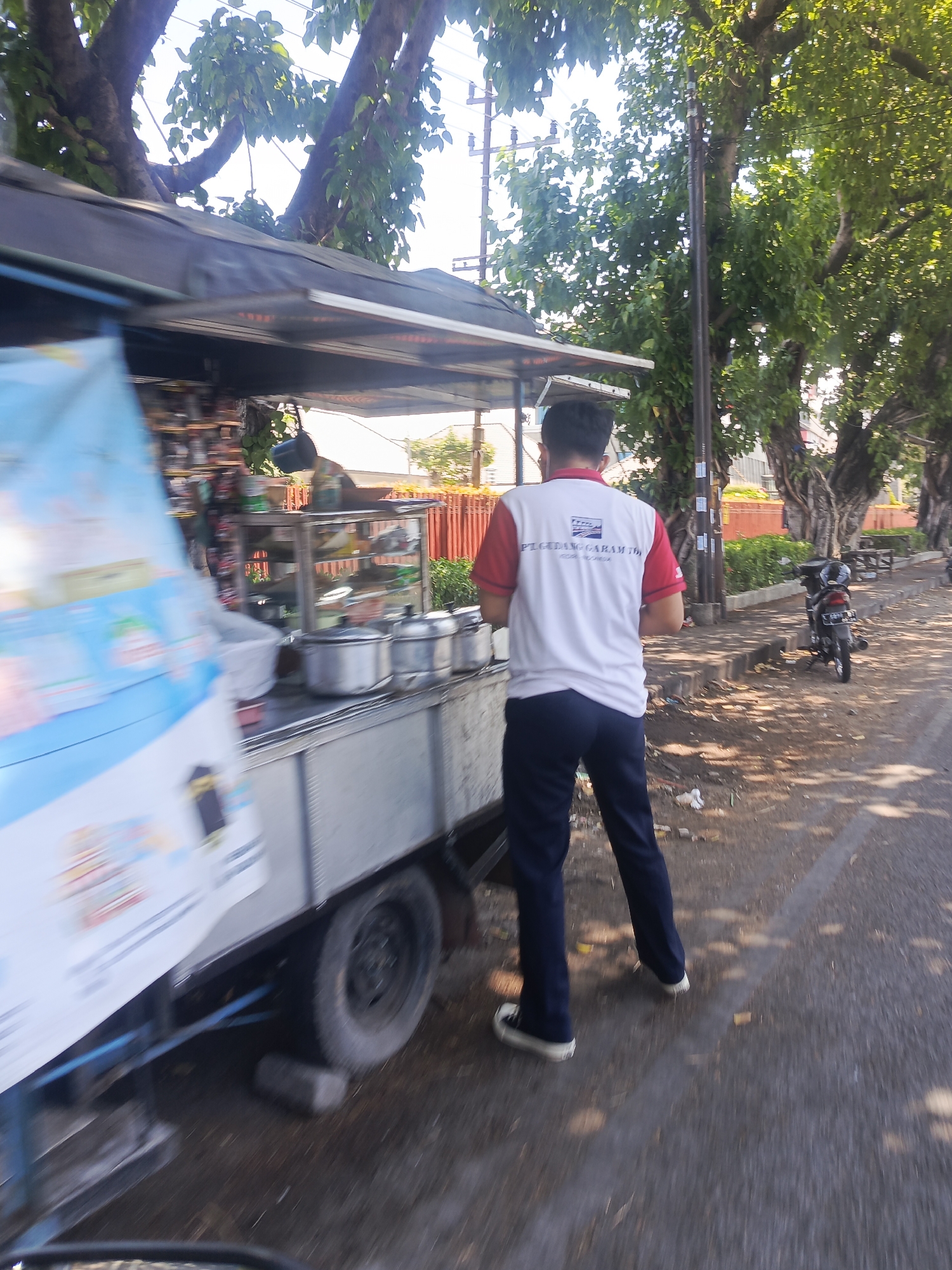 An exterior, eye-level, medium shot captures a street scene in Kedungrejo, Indonesia, on a sunny day with dappled light. In the foreground, a man stands facing away from the camera, wearing a white polo shirt with red sleeves and trim, and dark blue trousers. His shirt bears a logo and text which reads "PEGILAGAN GARAM TIRTA". He is positioned beside a food cart, which is filled with various containers and items. The cart has a dark canopy and a silver-colored side. Behind the cart, a street is visible with trees lining the sidewalk and a parked motorcycle. The ground is paved and appears somewhat wet in places. In the background, buildings and more trees are visible under a bright sky. The overall impression is one of an ordinary day in a bustling Indonesian town.