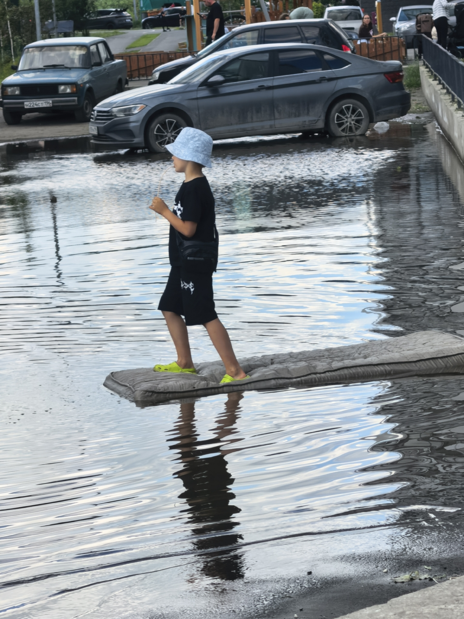 A young person wearing a light blue bucket hat, a black t-shirt, and black shorts walks across a flooded area. They are wearing bright yellow Crocs and carrying a black bag slung across their chest. The person is standing on a gray, rectangular object that appears to be floating or resting on the water's surface, possibly a cushion or mat. The water is calm with ripples reflecting the sky and surrounding objects. In the background, several cars are visible: a light blue sedan with a license plate that reads "H224CT 196", a dark gray sedan, and other vehicles further away. The scene suggests an outdoor environment, possibly a street or parking lot that has been flooded. The lighting indicates it is daytime, and the sky's reflection suggests it might be a partly cloudy day. There are no obvious signs of distress or urgency, suggesting a playful or resourceful approach to navigating the flooded conditions. The overall impression is one of overcoming a common obstacle in a creative way.