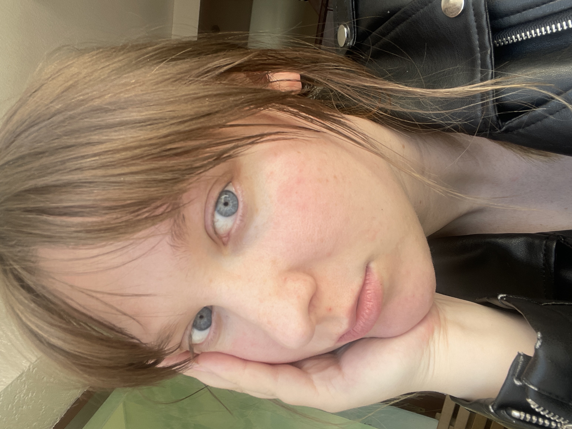 A close-up, slightly angled portrait of a fair-skinned person with light brown hair and blue eyes. The person's face rests on their hand. The person is wearing a black leather jacket. The background is indistinct. The image does not contain location information relating to Noviy, Russia.