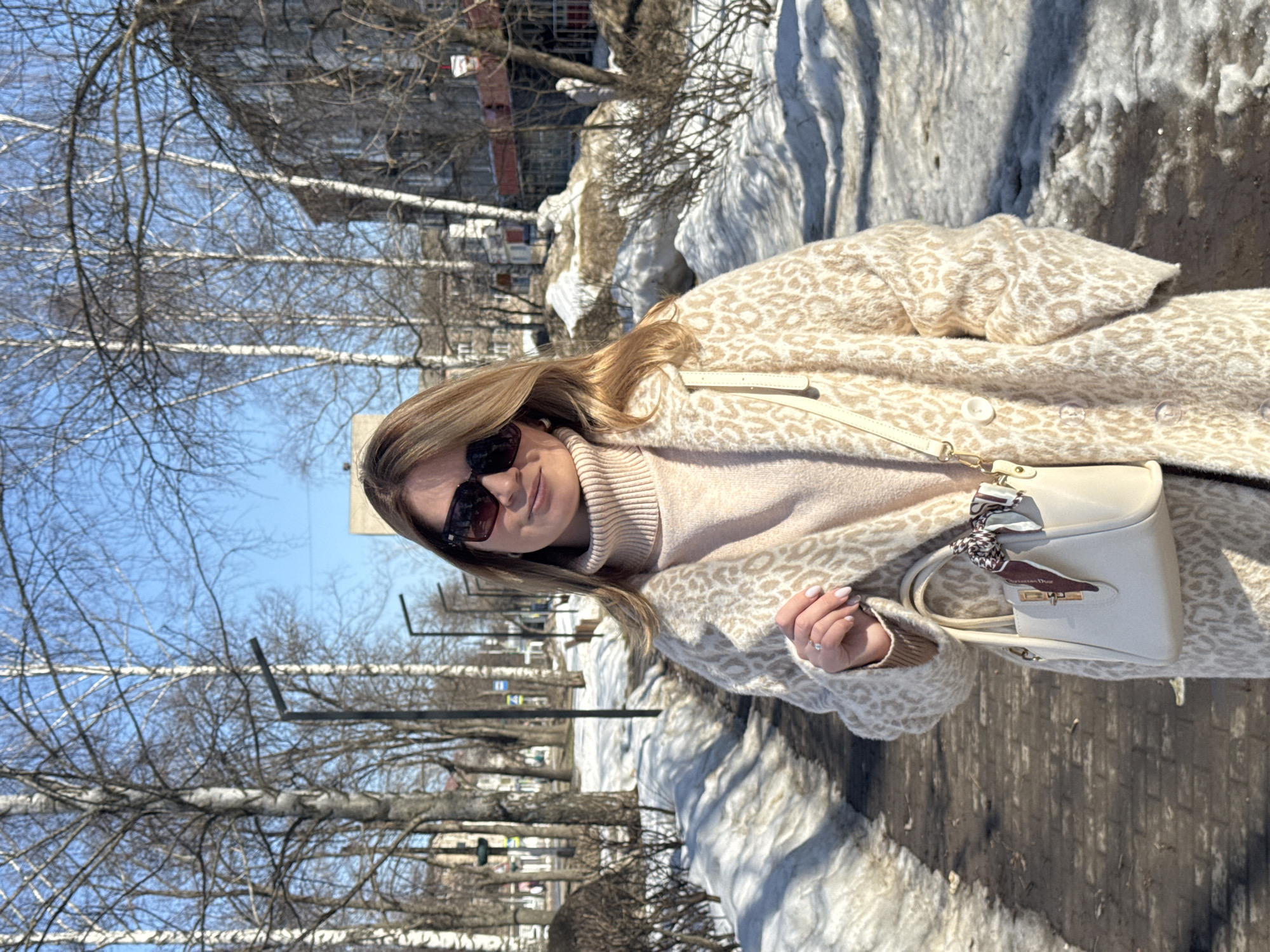 A woman in sunglasses and a patterned coat stands on a sidewalk next to a snow-covered area. She is wearing a light pink sweater and holding a small, light-colored handbag. The background shows a row of trees without leaves, a street with lamp posts, and buildings. The photo was taken in Chaikovsky, Russia, as determined by the provided location data.