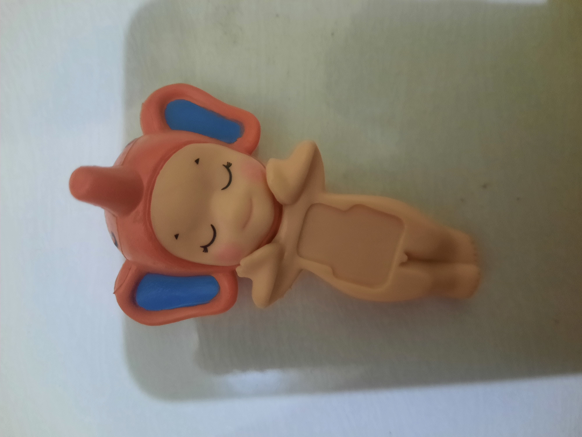 A small, pale pink figurine resembling an elephant stands on a light-colored, slightly textured surface. The figurine has large, blue-lined, orange elephant ears, closed eyes with curved black lines, and rosy cheeks. Its body is a smooth, skin-toned color, and it has small, raised arms held across its chest. The surface on which the figurine rests is lit from the left, casting a soft shadow to the right of the object. The background is a uniform, neutral color. There is no indication of the city of Lhokseumawe, Indonesia, or any urban environment in this image.