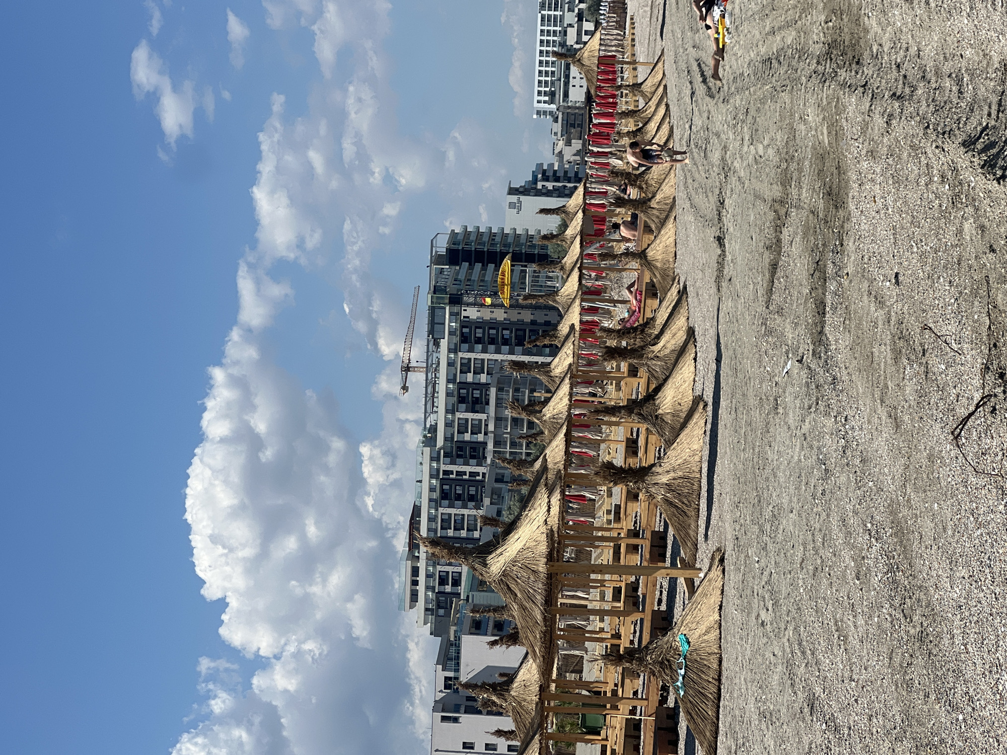The image shows a beach in Năvodari, Romania, with a row of thatched umbrellas and lounge chairs. In the background, modern apartment buildings line the coast, with a construction crane visible among them. A few people are scattered on the beach, with one person standing near the umbrellas and another lying down on a lounge chair. The sky is blue with scattered white clouds. The beach appears to be made of sand and small pebbles.