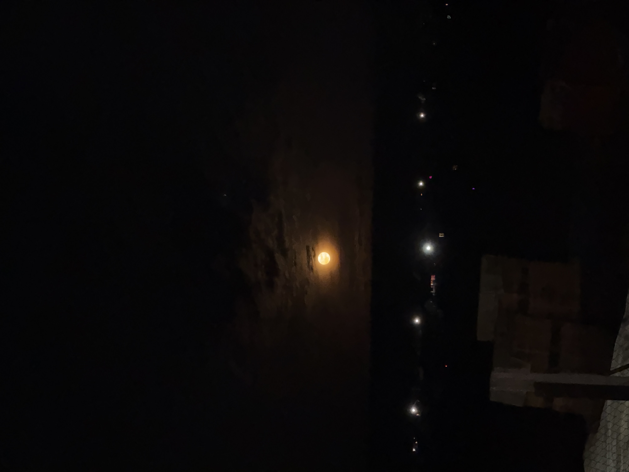 The image depicts a night scene in Горный, Russia. A full moon is partially obscured by clouds in the center of the frame. To the right, a string of lights descends vertically, likely from a building or structure. Adjacent to the lights, a stacked arrangement of boxes or containers is partially visible. The overall environment is dark.