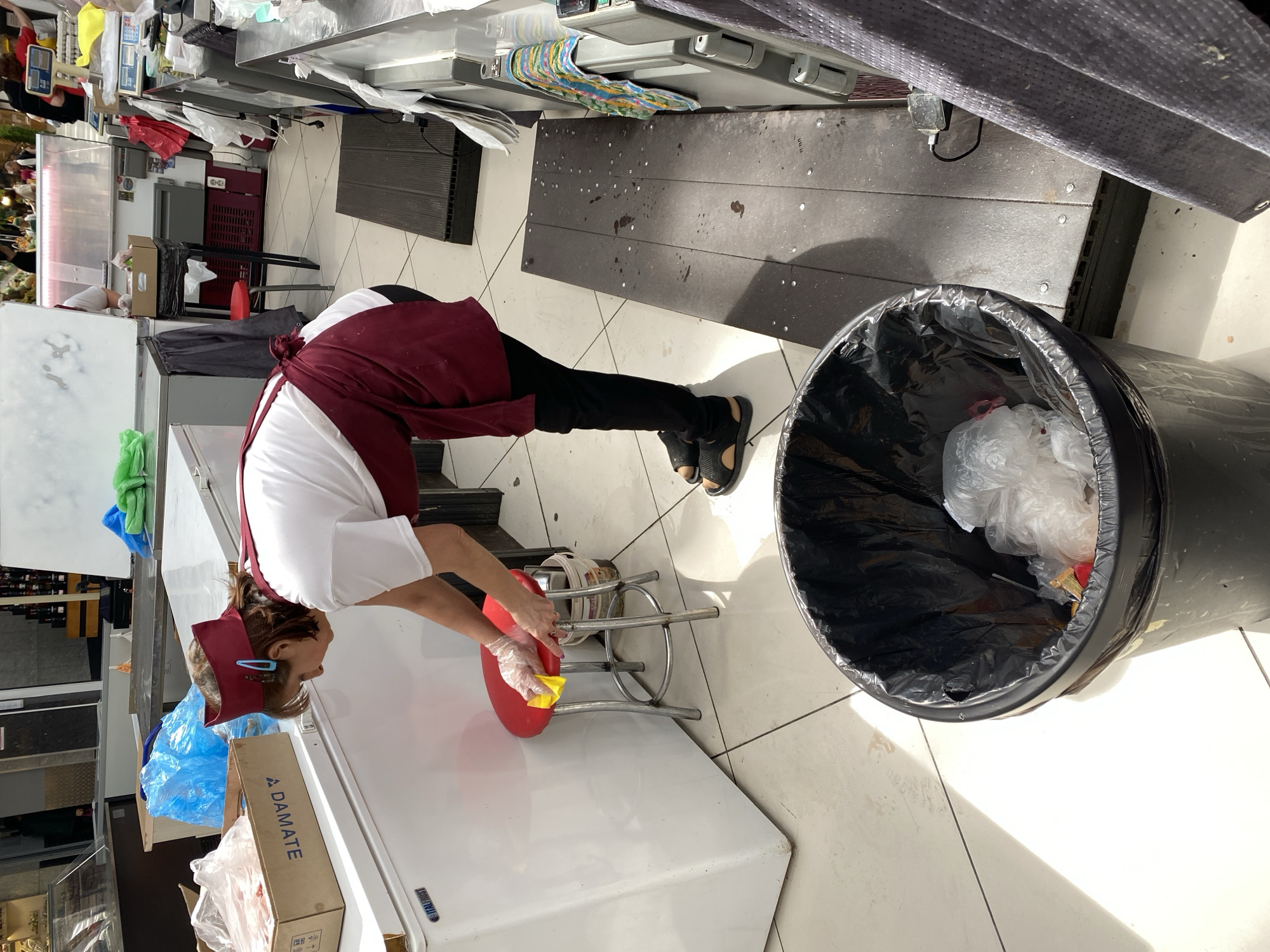 A female employee wearing a burgundy apron and a red headband is cleaning a red stool with a yellow cloth in what appears to be a food market in Novaya Usman', Russia. She is wearing gloves. The environment includes a large white freezer unit, a trash can with a black liner filled with plastic bags, and various market stalls and equipment in the background, including a scale and display cases. The floor is tiled and appears to be a light color.