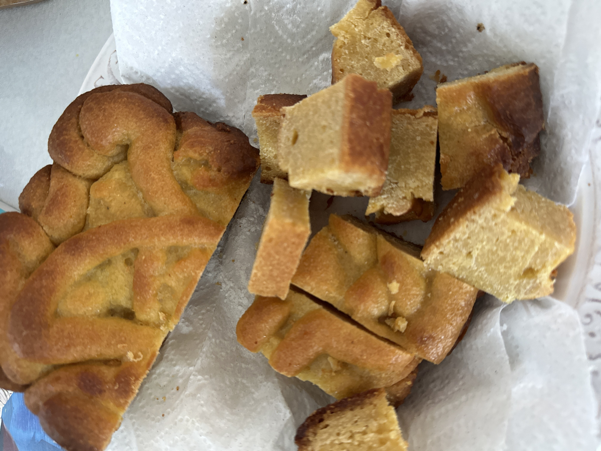 The image shows a close-up view of a baked good, likely a cake or bread, that has been cut into several pieces. A portion of the whole item is visible at the top, with a braided or latticed crust. The remaining pieces are scattered on a white paper napkin, some of which are also cut into smaller cubes. The baked good appears to be golden brown with a slightly dense texture. The background is blurred and shows hints of blue and white. The image does not contain any information that would allow for the determination of its location in the city of Nevinnomyssk, Russia.