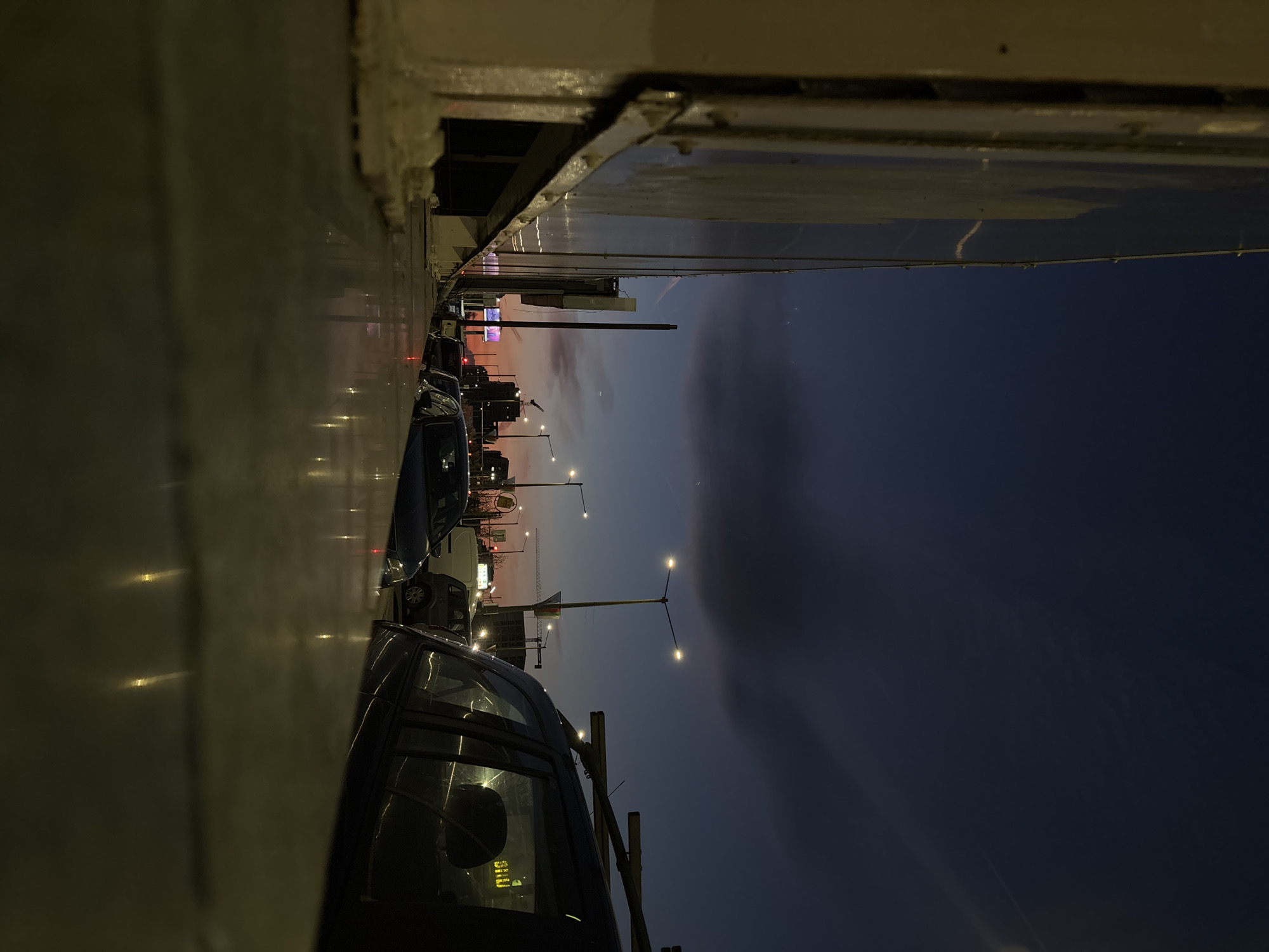 The image shows a street scene in Sofia, Bulgaria at dusk, viewed from a high angle. A row of parked cars lines the left side of the street. Streetlights illuminate the road, and building lights are visible in the distance. The sky transitions from a pale orange near the horizon to a dark blue above. The perspective is partially obscured by a concrete structure at the top and left of the frame.