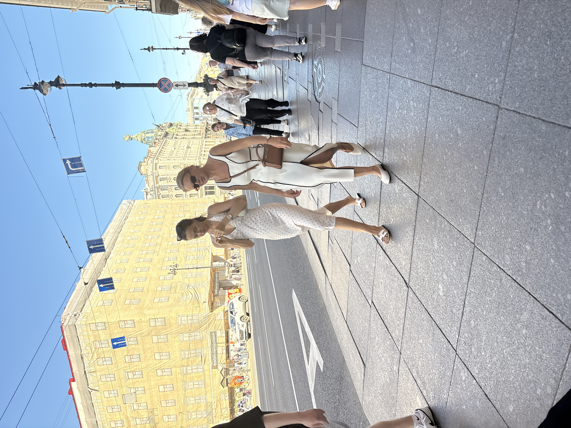 On a sunny day in Saint Petersburg, Russia, two women walk along a wide sidewalk paved with large granite tiles. The woman in the foreground, on the right, wears a sleeveless white dress with black trim and a brown crossbody bag. She has blonde hair and is wearing sunglasses. The woman to her left wears a white polka dot dress and white sandals. Behind them, a street with cars and pedestrian traffic is visible, with buildings lining the other side. The building directly behind the women is covered in a large yellow tarp, suggesting ongoing construction or renovation. The sky is clear and blue.