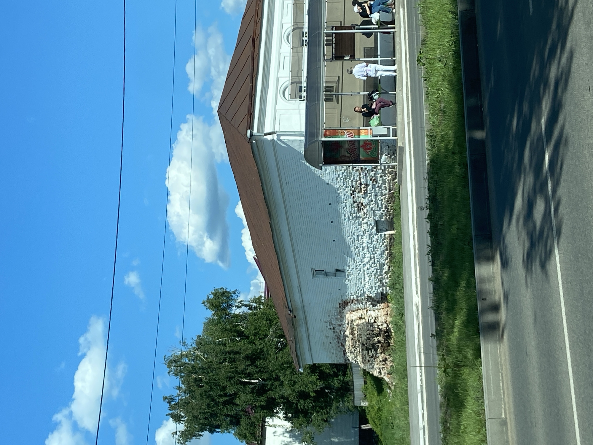 The image captures a bright daytime scene in Chistopol, Russia, featuring a two-story building alongside a paved road under a clear blue sky with scattered white clouds. The photograph is oriented vertically, showing the building on the left and the road on the right.
The prominent building has a brown sloped roof and light-colored walls. Its lower section displays a textured, possibly distressed, brick or stone facade with some areas showing exposed brickwork. The upper story features arched windows. An extended awning or covered area is attached to the building's front, beneath which several people are gathered.
Five individuals are visible in this covered area. One person, dressed in a white top and light pants, stands facing right. Four other people are seated: one in a dark top and light pants, two further back, and one closer to the foreground in a dark top and maroon pants, with a green bag resting beside them. They appear to be waiting or resting, possibly at a bus stop or outside a commercial establishment.
A narrow strip of green grass separates the building and its sidewalk from the paved road. The road has a solid white line marking its edge and a dashed white line further towards the center. Strong, dark shadows of trees are cast across the asphalt, indicative of bright sunlight. Overhead power lines stretch across the sky. Green foliage from trees is visible behind and beside the building.
A colorful sign with Cyrillic text, possibly "ЖИВАЯ" (Zhivaia), and a green logo is visible in a window under the awning, likely an advertisement.