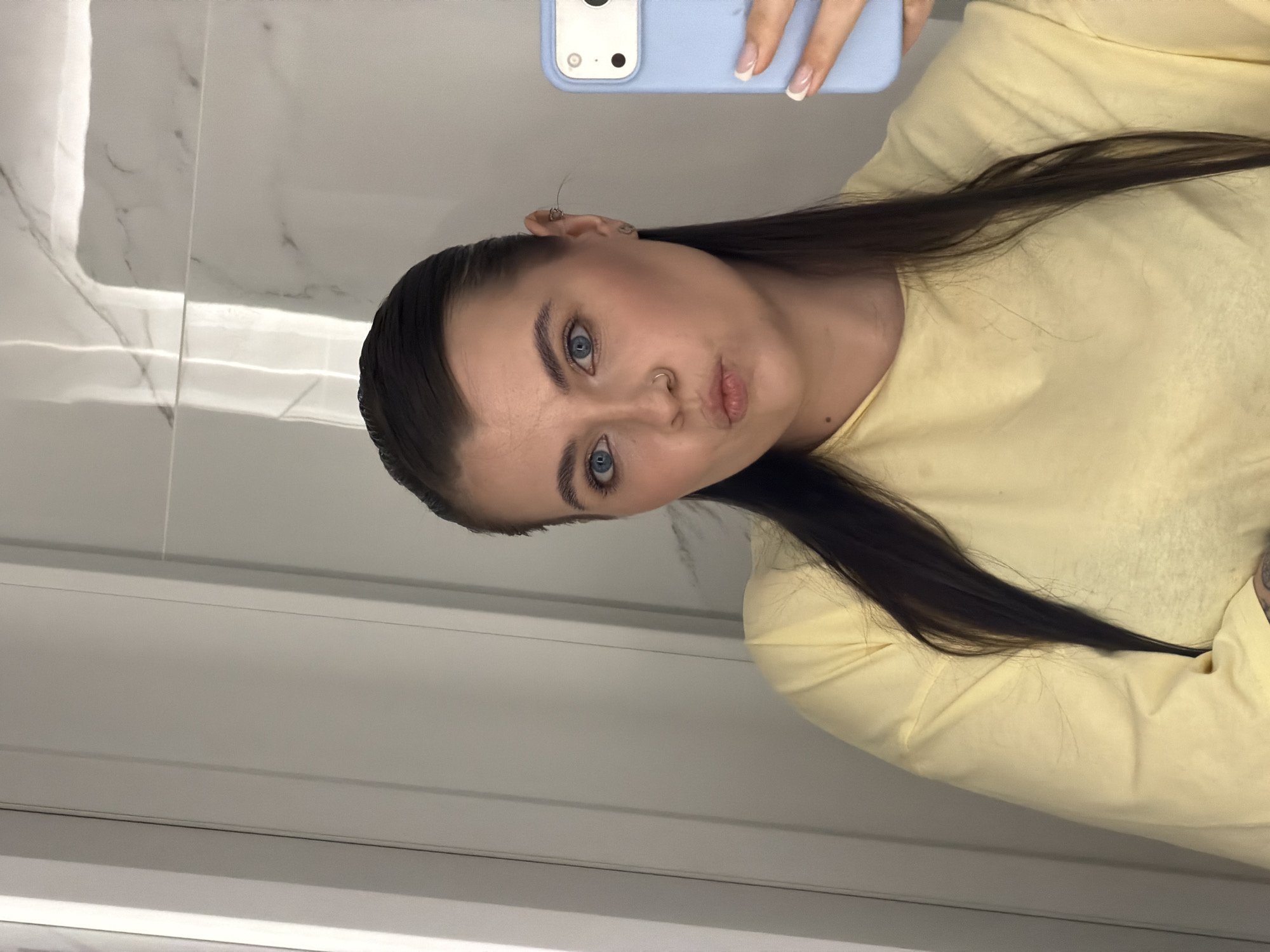 A mirror selfie captures a young woman in an indoor setting in Chaikovsky, Russia. She has long, dark hair styled back, prominent blue eyes, and defined eyebrows. Her lips are playfully puckered, and her gaze is directed towards the camera. She is wearing a light yellow long-sleeved top. Notable accessories include a small silver nose ring and subtle ear piercings on her visible left ear.
The woman is holding a light blue smartphone with her left hand, which features French-tipped fingernails. The phone's case and camera module (two dark circles) are visible in the upper portion of the reflection. The background behind her in the mirror appears to be a light-colored, possibly white or off-white, tiled wall with subtle gray veining, resembling marble. Horizontal lines below the main reflective surface suggest architectural paneling or additional tiling. The lighting is artificial, characteristic of an indoor environment like a bathroom or changing room. No text is visible in the image.