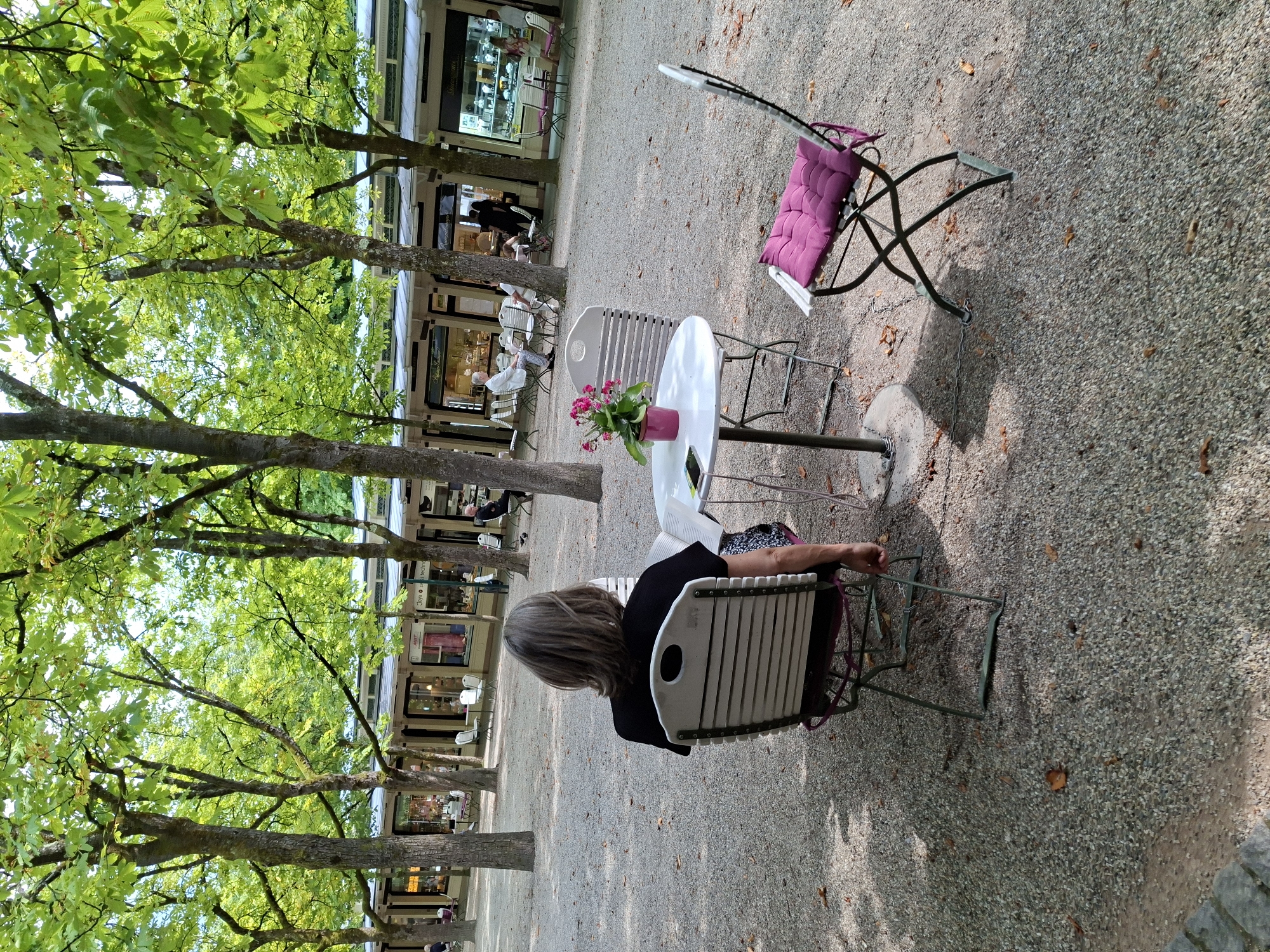 The image depicts a serene outdoor seating area in Baden-Baden, Germany, likely within the Lichtentaler Allee, a renowned park. In the foreground, a woman with grey hair sits with her back to the camera, facing a small round table with a pink potted flower and a mobile phone. Two chairs are positioned around the table, one occupied by the woman, the other empty with a purple cushion. Beyond this central scene, a row of shops or cafes with glass fronts is visible, shaded by mature trees with lush green foliage. Several other patrons are seated at tables outside these establishments, enjoying the tranquil ambiance. The ground is covered with small grey gravel and scattered autumn leaves, suggesting a pleasant daytime setting in either spring or fall.