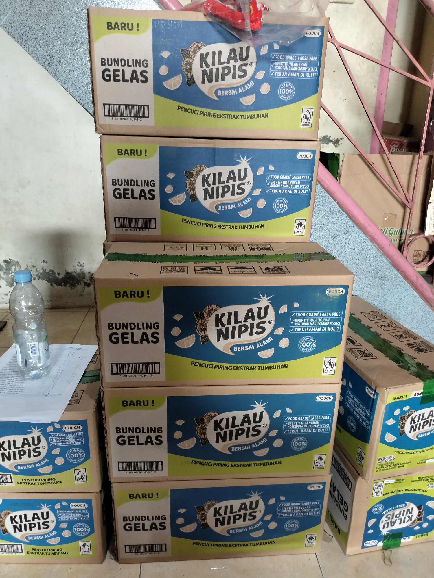 The image shows a stack of cardboard boxes, likely containing dish soap. The boxes are blue and yellow with the brand name "KILAU NIPIS" prominently displayed. Text on the boxes indicates "BUNDLING GELAS" (glass bundle), "PENGISI PIRING EKSTRAK TUMBUHAN" (dish soap with plant extract), and claims it is "FOOD GRADE' LABSA FREE" (food grade and chemical free), "EFEKTIF HILANGKAN KOTORAN & BAU CUKUP 1X CUC" (effectively removes dirt and odor with just one wash), and "TERUJI AMAN DI KULIT" (tested safe for skin). Some boxes also feature a "HALAL" certification logo and a 100% "KEKUATAN" (strength) seal. The boxes are stacked in a slightly haphazard manner, with some leaning against each other. To the left of the boxes, a clear plastic water bottle with a blue cap rests on a white document. The background shows a textured wall with some discoloration and a pink metal structure. The lighting suggests an indoor environment, possibly a storage area or a shop. While the prompt asks for the location in the city of Langsa, Indonesia, there are no visual cues in the image that specifically identify this location.