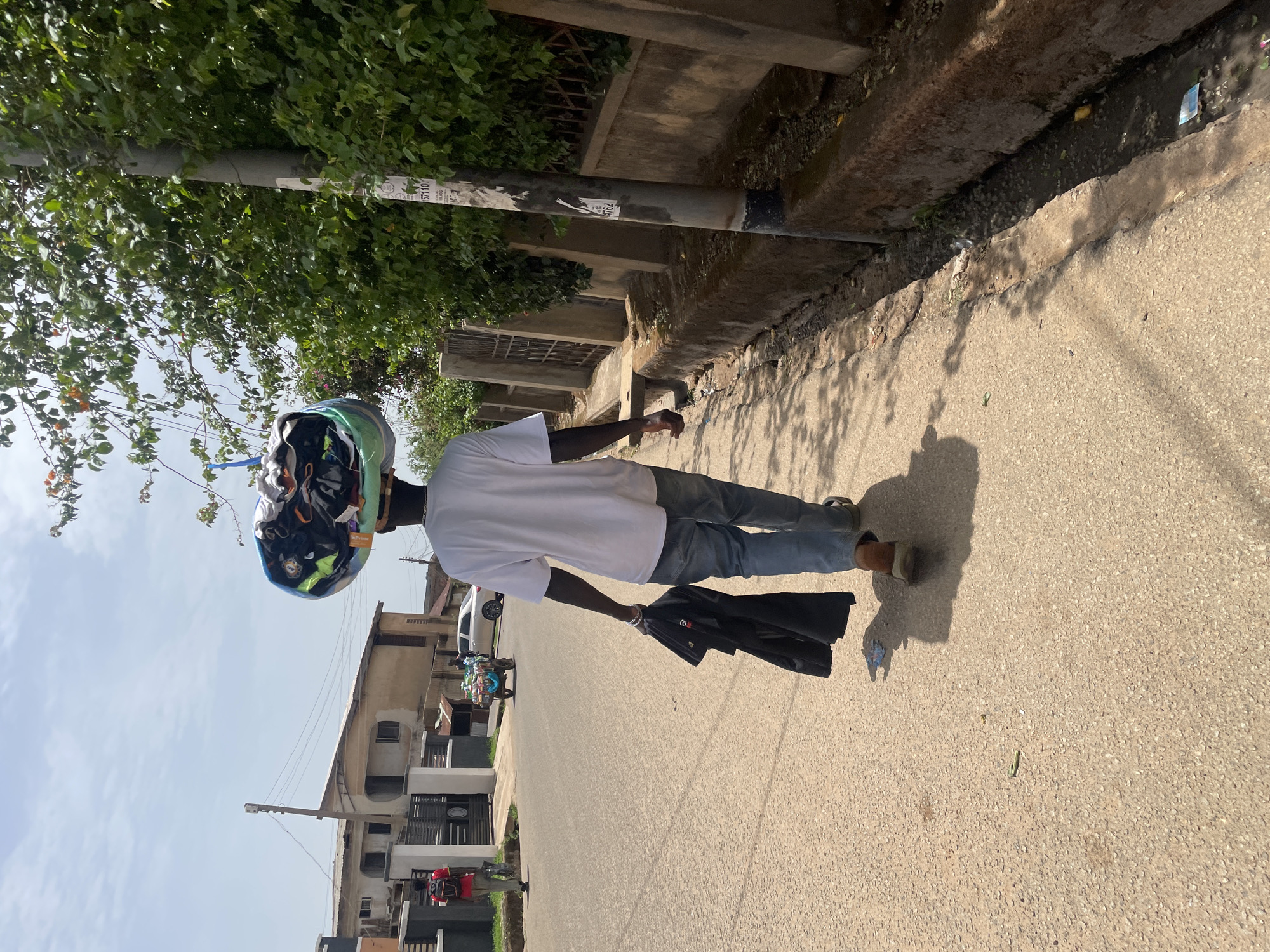 The image depicts a man walking on a paved street in a residential area of Jos, Nigeria. He wears a white t-shirt, blue jeans, and sandals, and carries a folded black umbrella in his left hand. On his head, he has a bundled assortment of items secured with straps. The street is bordered by buildings on one side and a raised concrete edge with a tree and foliage on the other. A utility pole is visible along the edge of the street. A vehicle and other individuals are visible in the distance. The sky is partly cloudy.
