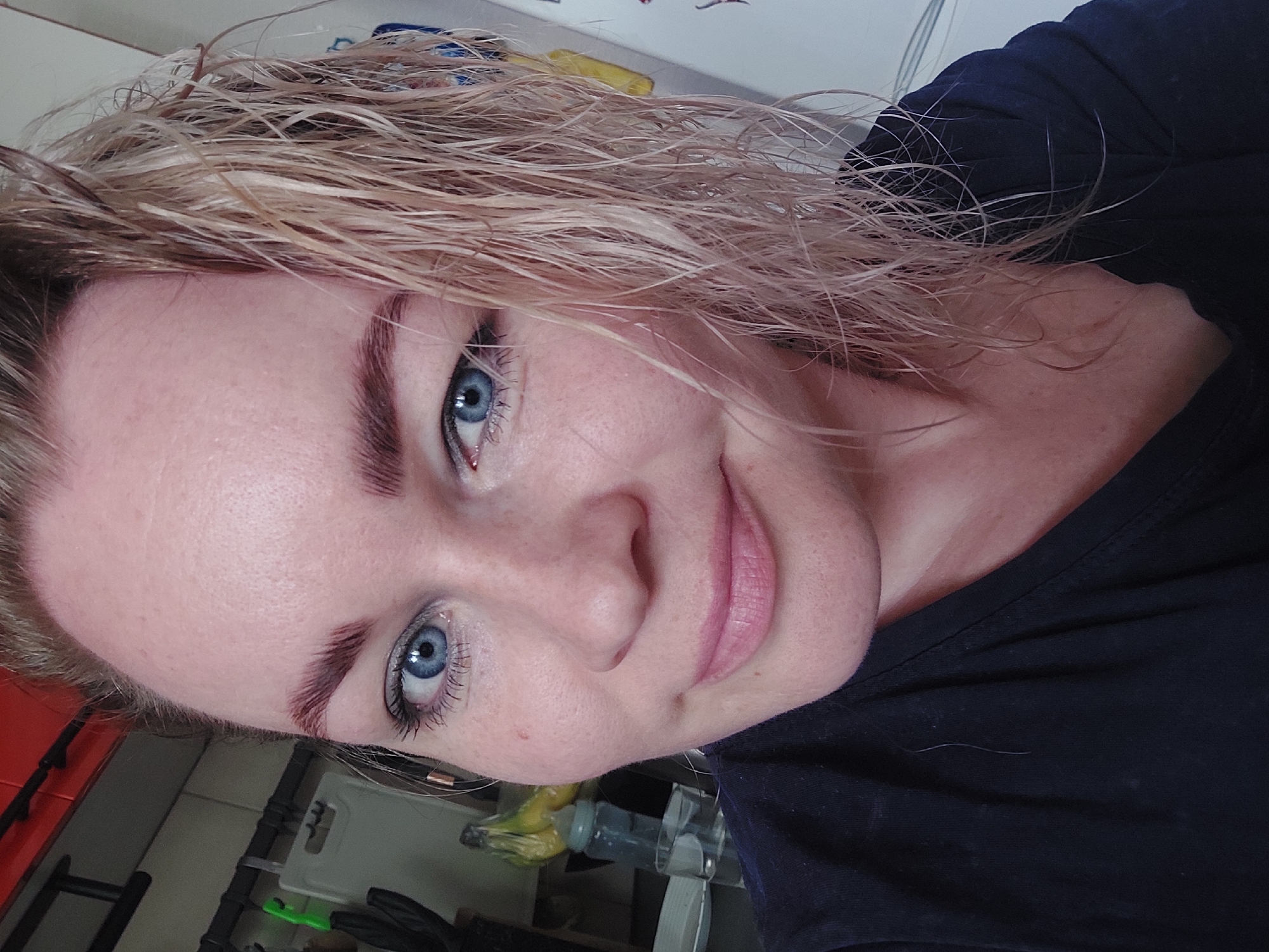 A close-up, waist-up photograph shows a fair-skinned woman with blue eyes and wet, blonde hair. Her hair is styled to fall over the right side of her face. She is wearing a dark blue V-neck shirt and has applied makeup, including mascara and lip gloss. The background is blurred, but it appears to be an indoor setting with what looks like kitchen cabinetry and a refrigerator visible. The lighting suggests it is daytime. The image was taken in Chaikovsky, Russia.