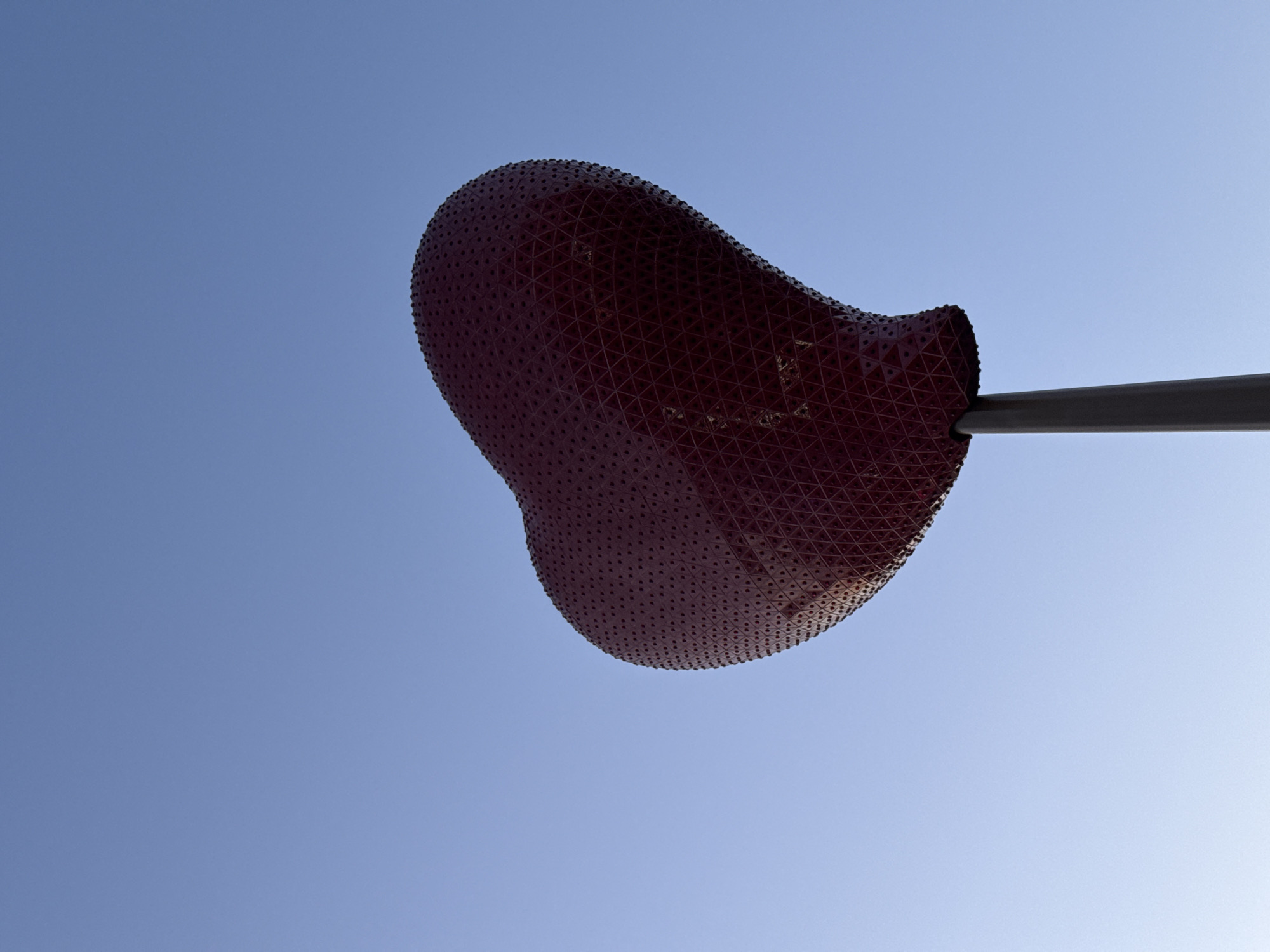 The image features a low-angle view of a maroon-colored, bean-shaped sculpture against a light blue sky. The sculpture has a perforated surface with a triangular pattern visible in its structure. A gray pole supports the sculpture from its right side. The sculpture is located in Paris, France. The overall impression is that of a modern art installation viewed on a clear, sunny day.
